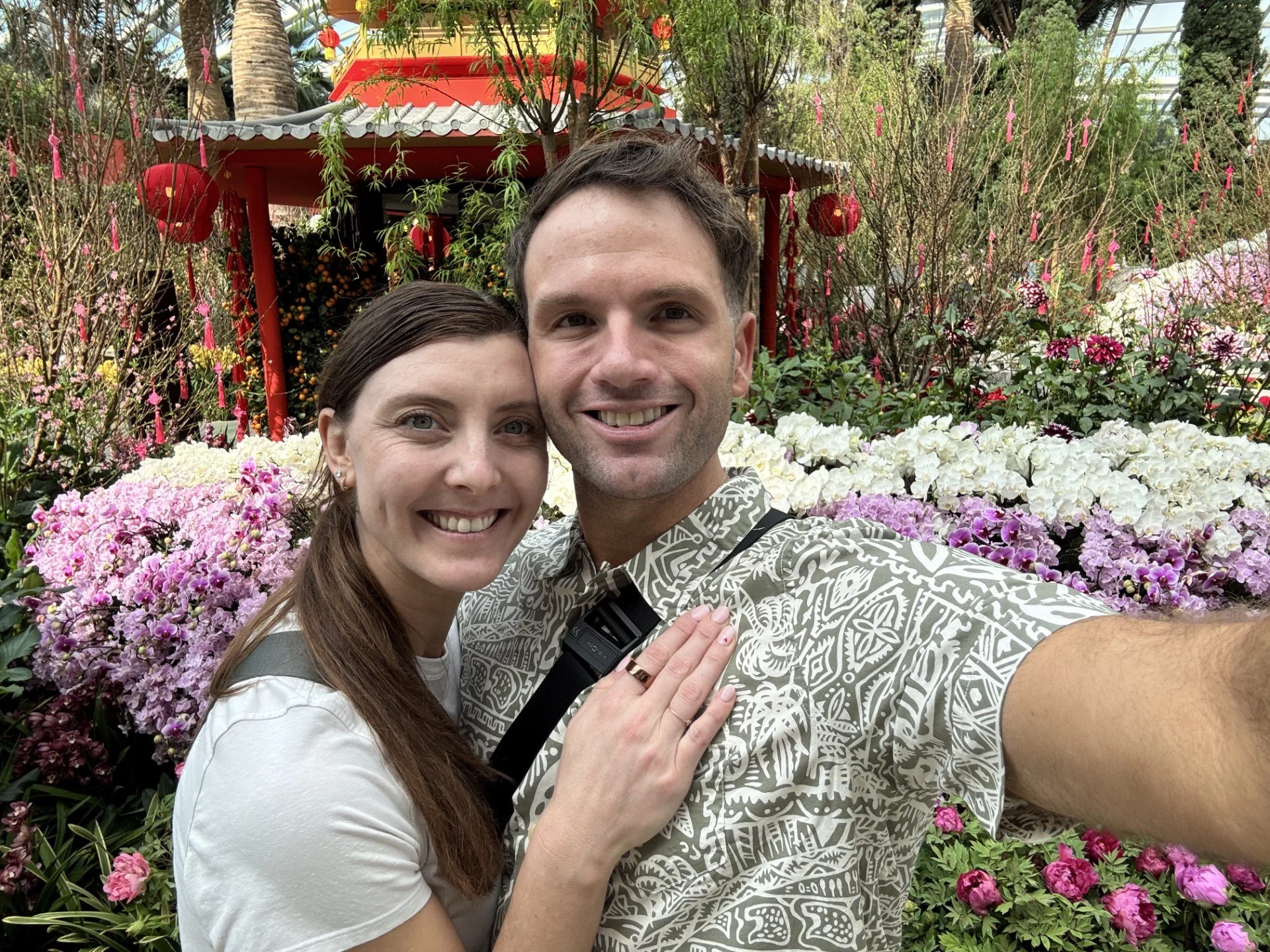 A smiling couple taking a selfie in a lush garden filled with colorful flowers and greenery, with a red pavilion in the background.