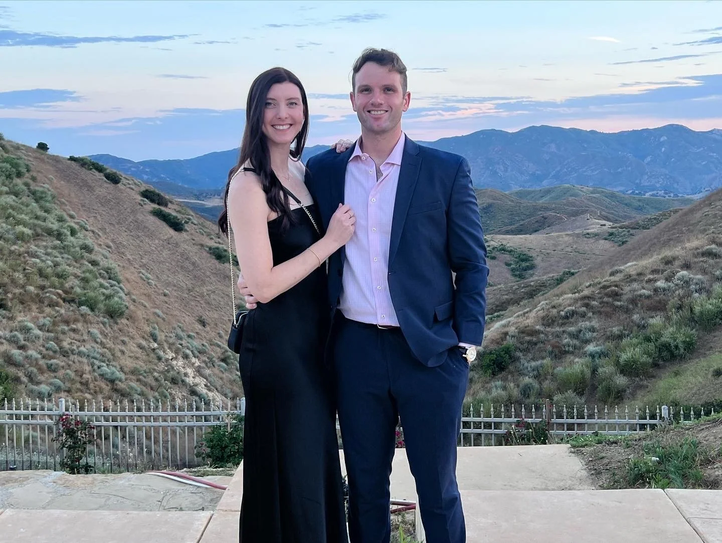 A smiling young couple dressed in formal attire standing close together outdoors, with rolling hills and mountains in the background at dusk.