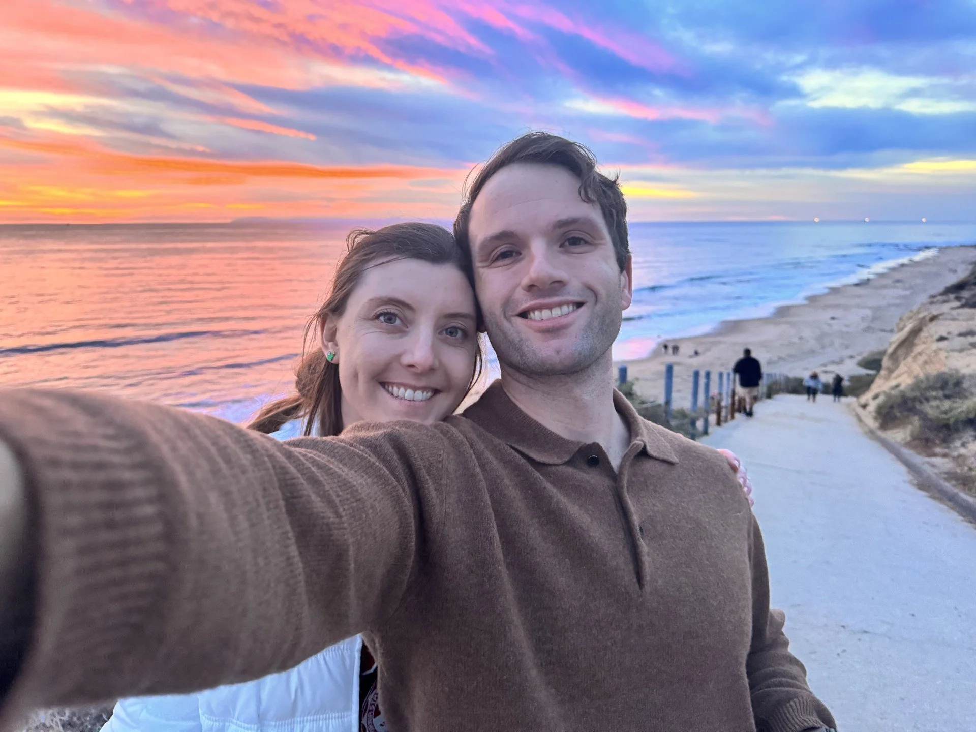 A smiling couple taking a selfie on a beach at sunset with colorful sky, ocean, and a pathway leading down to the beach in the background.