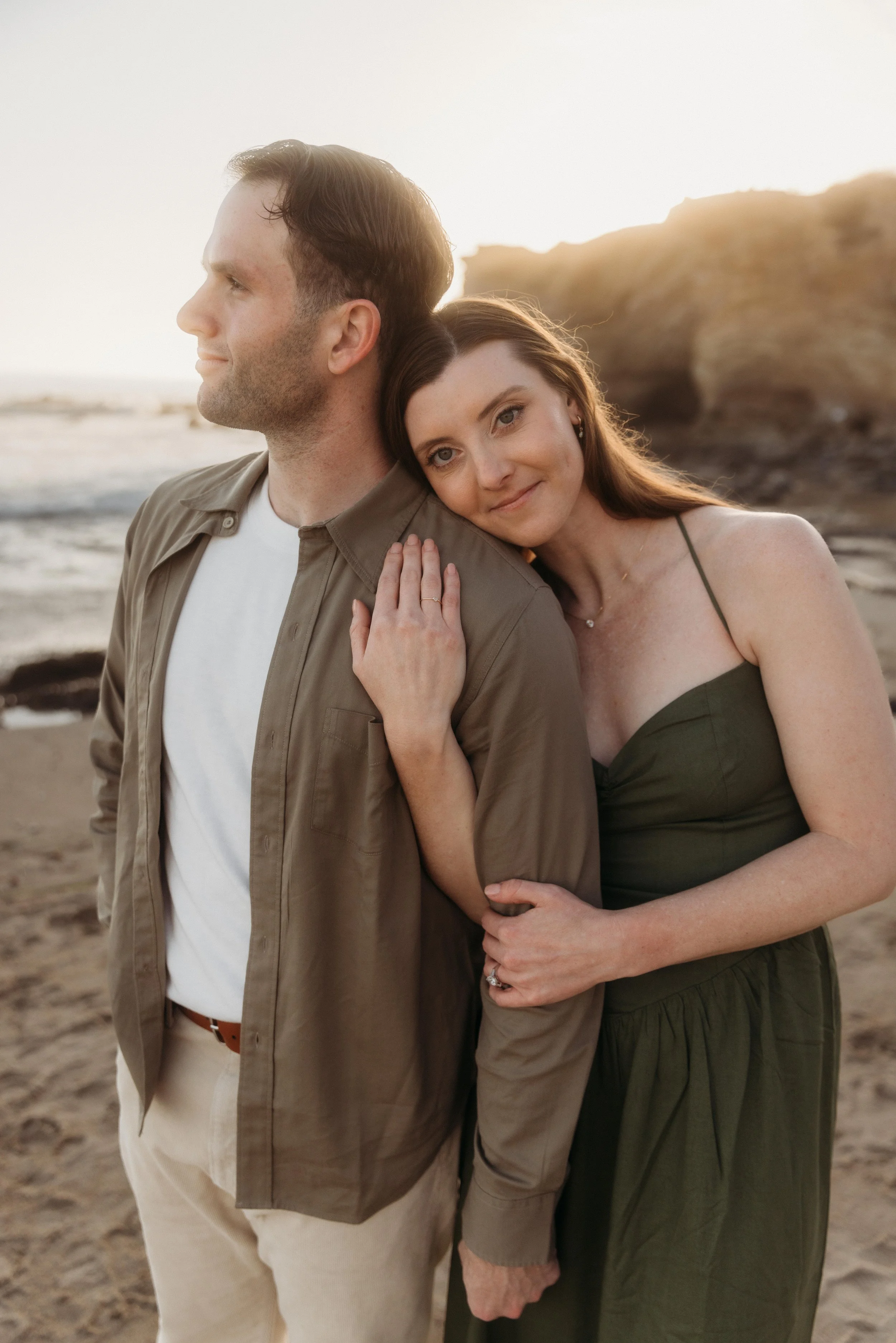 A woman with brown hair embracing a man at the beach during sunset, with rocks and ocean in the background.