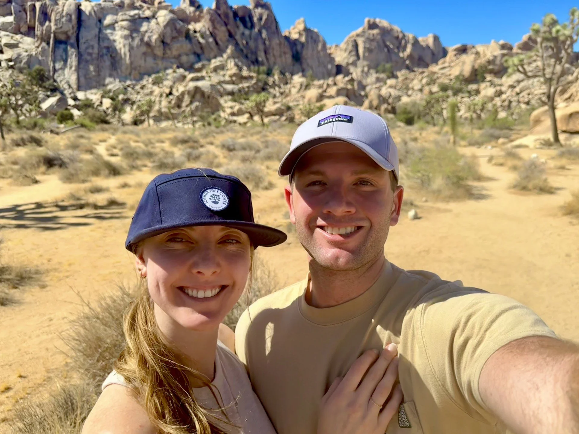 A smiling young couple taking a selfie outdoors in a desert landscape with rocky mountains and sparse vegetation in the background. They are wearing casual clothing and caps.