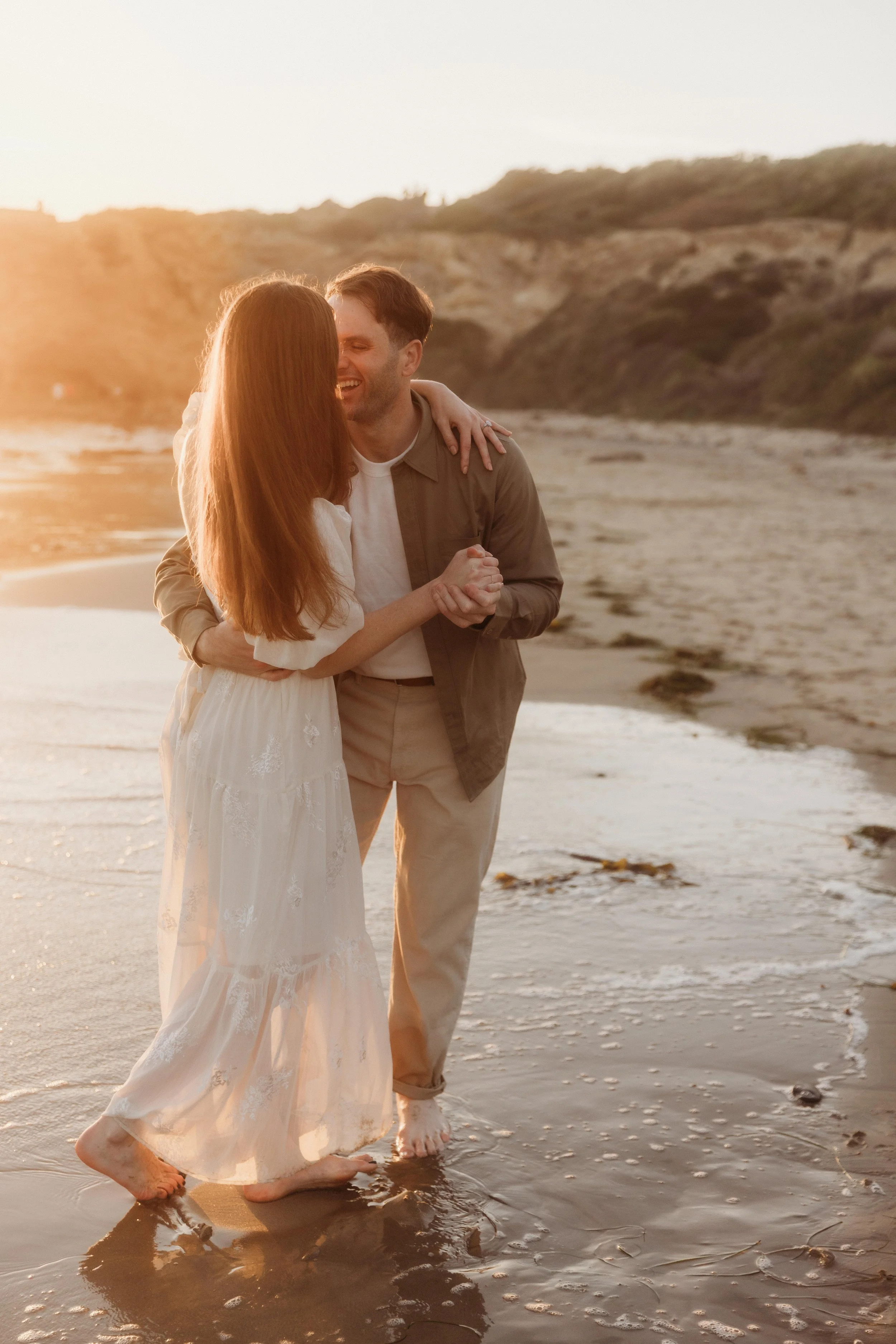 A couple standing barefoot on a beach at sunset, embracing and smiling at each other.