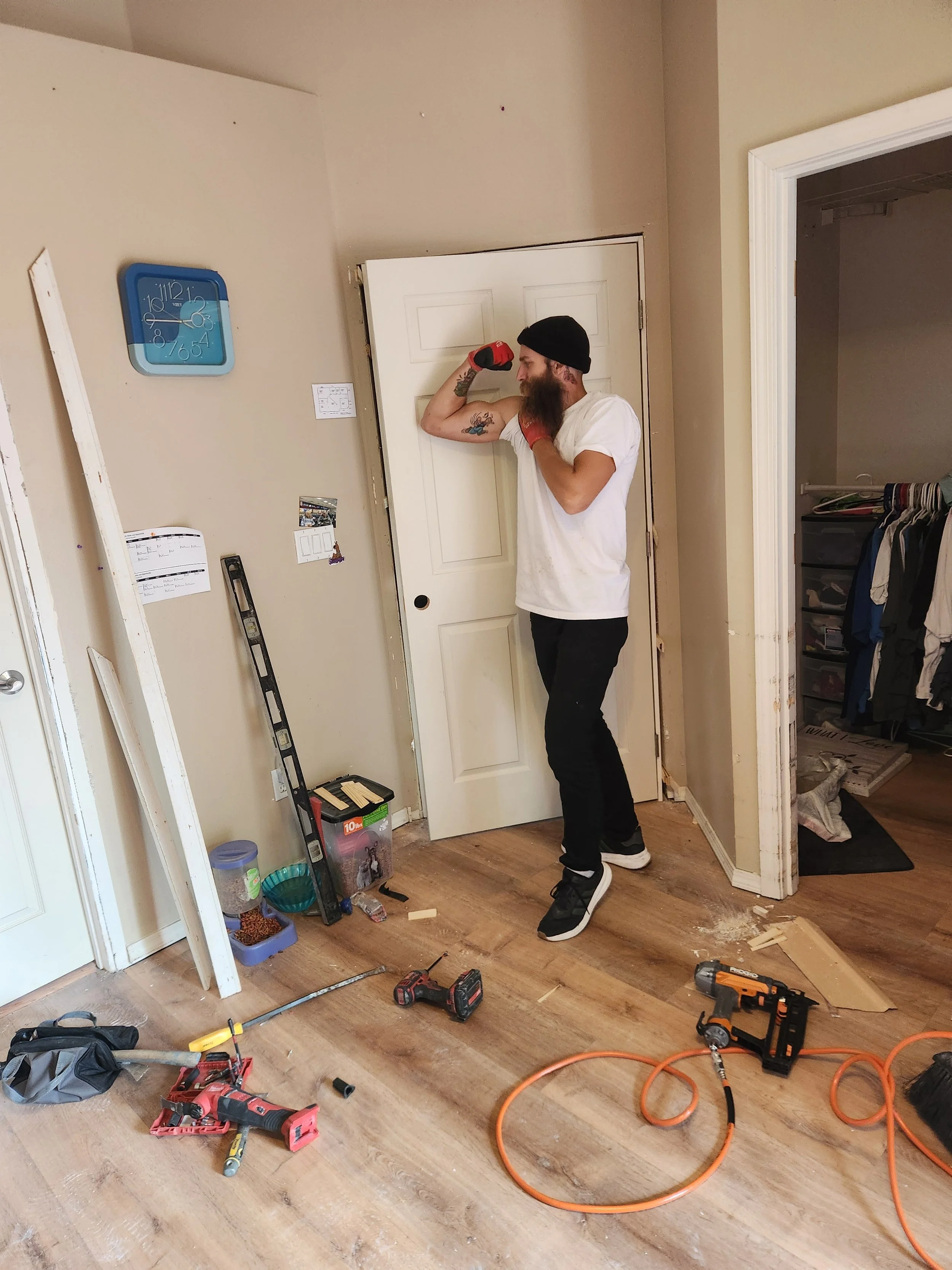 A man with a beard, wearing a black beanie, white T-shirt, and black pants, is posing playfully with a fake bicep curl gesture in a room under renovation. The room has tools on the floor, and a partially open closet with clothes inside.