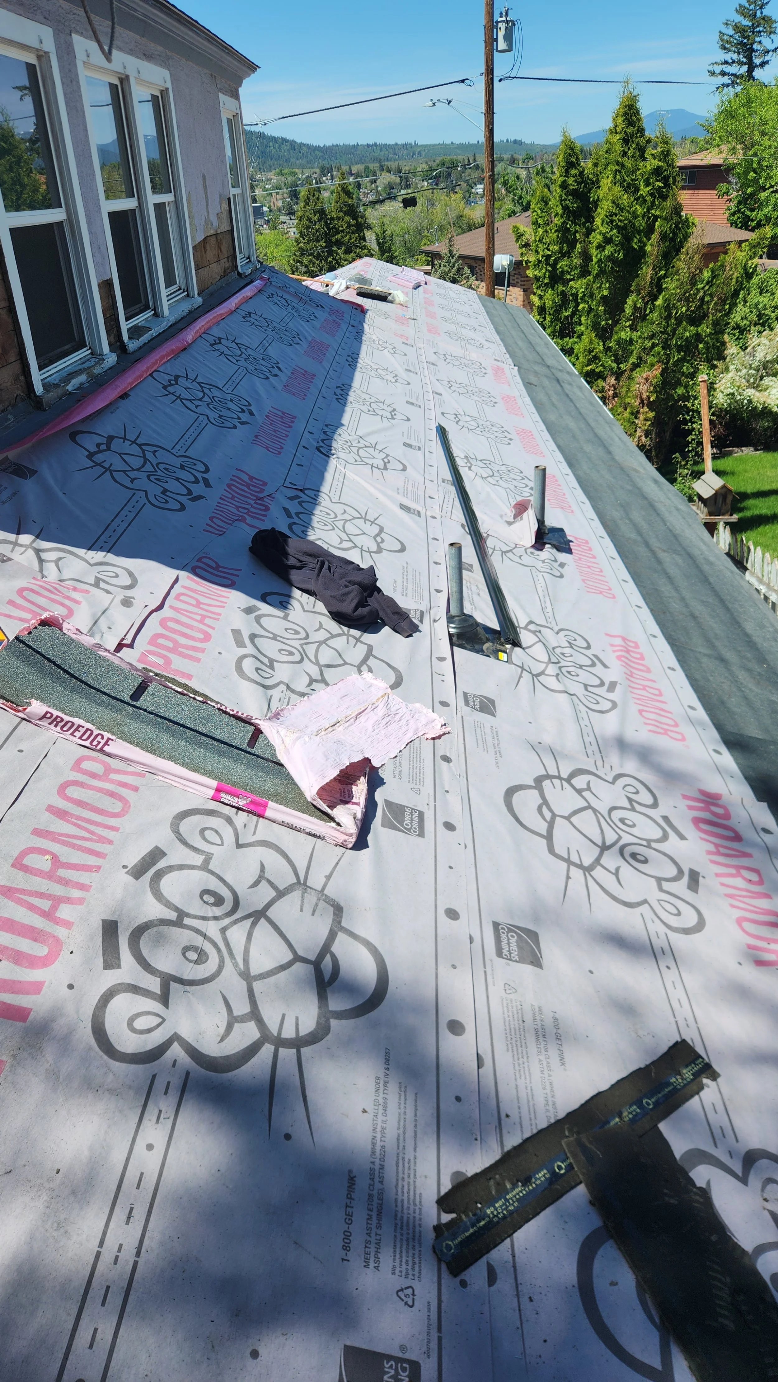 View of a roof under renovation with new roof membrane installed, construction tools and materials, and a residential neighborhood with trees and mountains in the background.