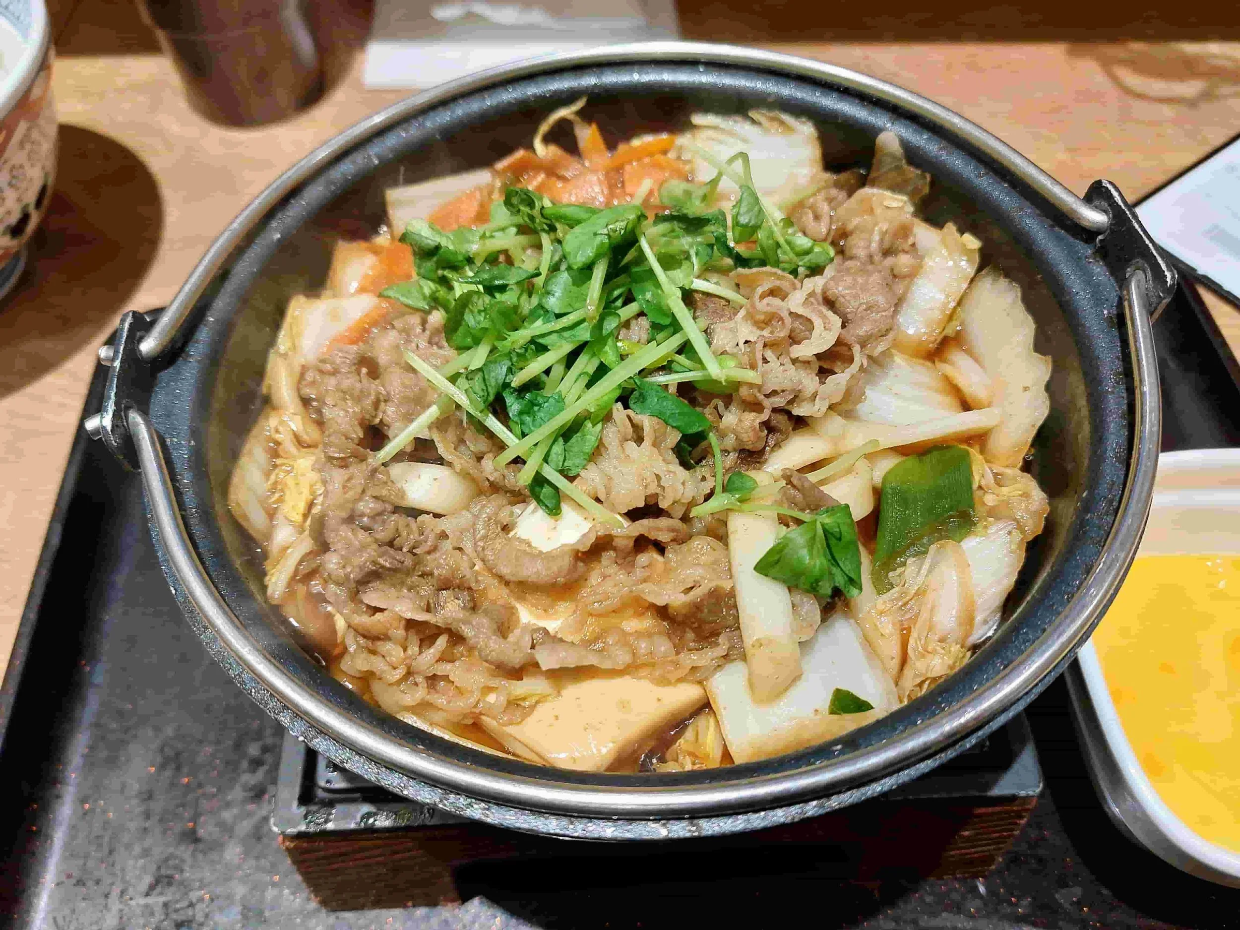 Sukiyaki hot pot with thinly sliced beef, tofu, vegetables, and noodles simmering in savory broth, served with raw egg for dipping