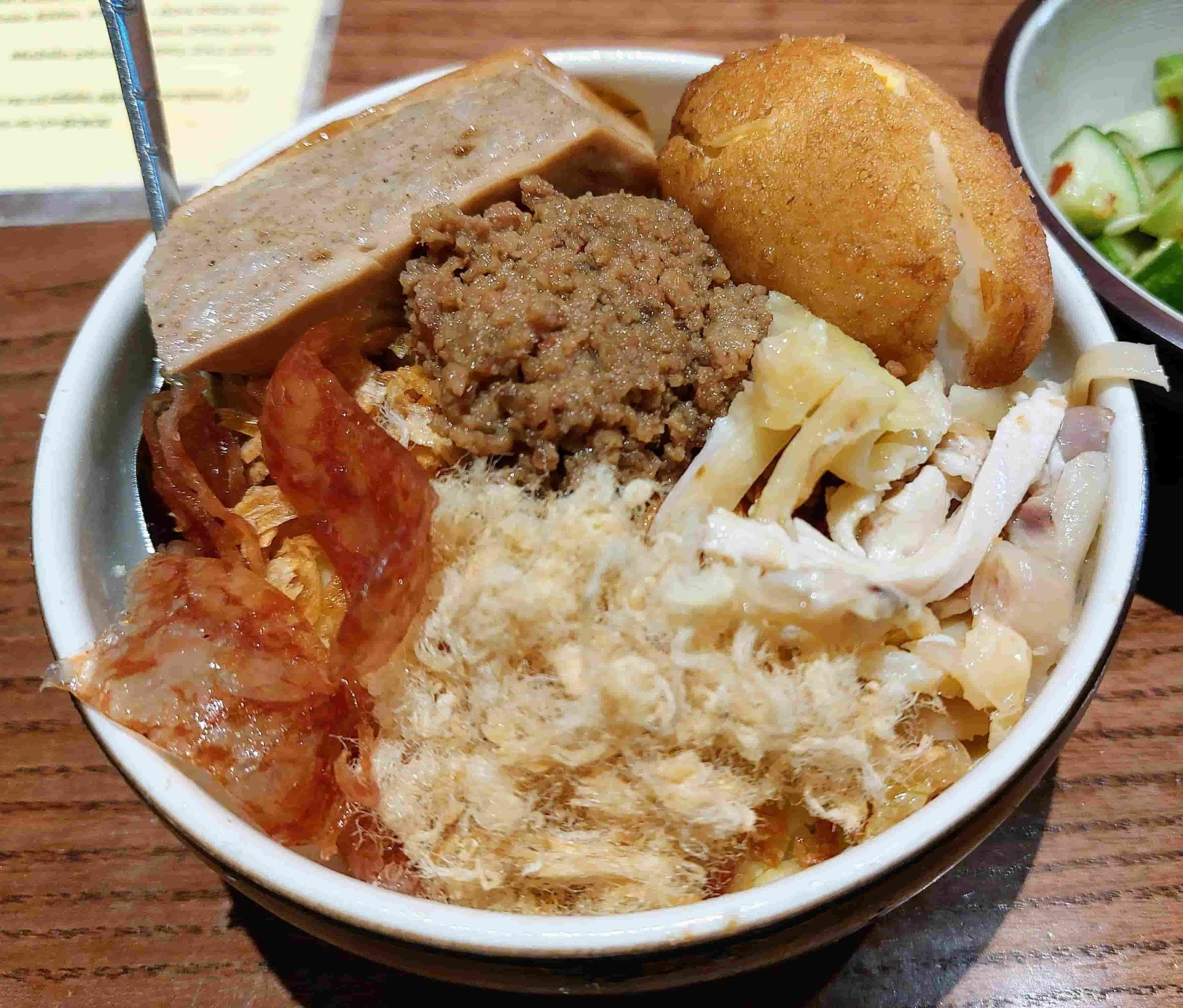 Bowl of Vietnamese xôi mặn (savory sticky rice) topped with shredded chicken, pork floss, Chinese sausage, pâté, cha lua (Vietnamese pork sausage), minced pork, and a halved deep-fried boiled egg. Served with a side of cucumber salad.