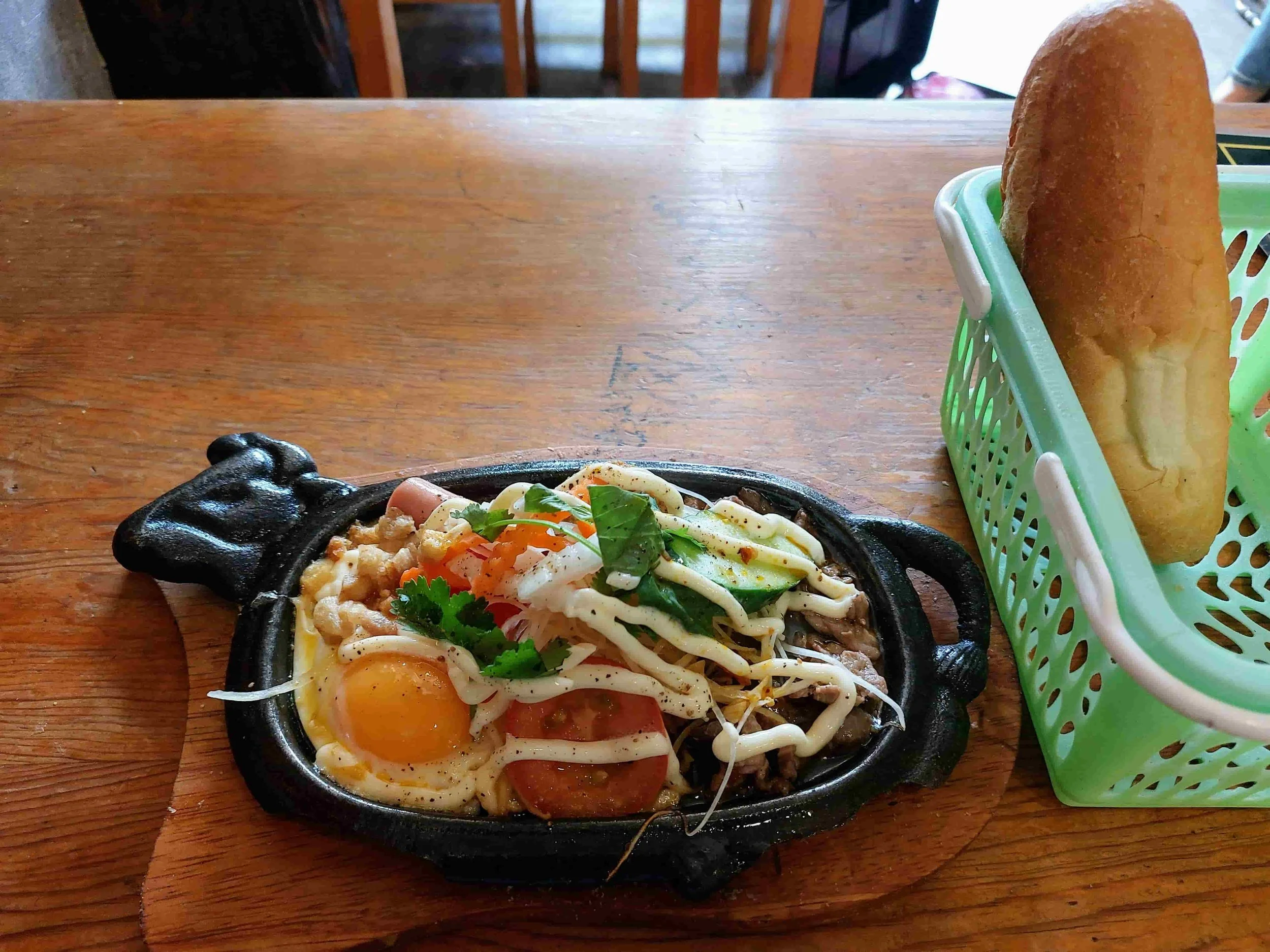 Vietnamese bò né served sizzling on a hot plate with thin slices of beef, a sunny-side-up egg, pâté, and a small baguette on the side.