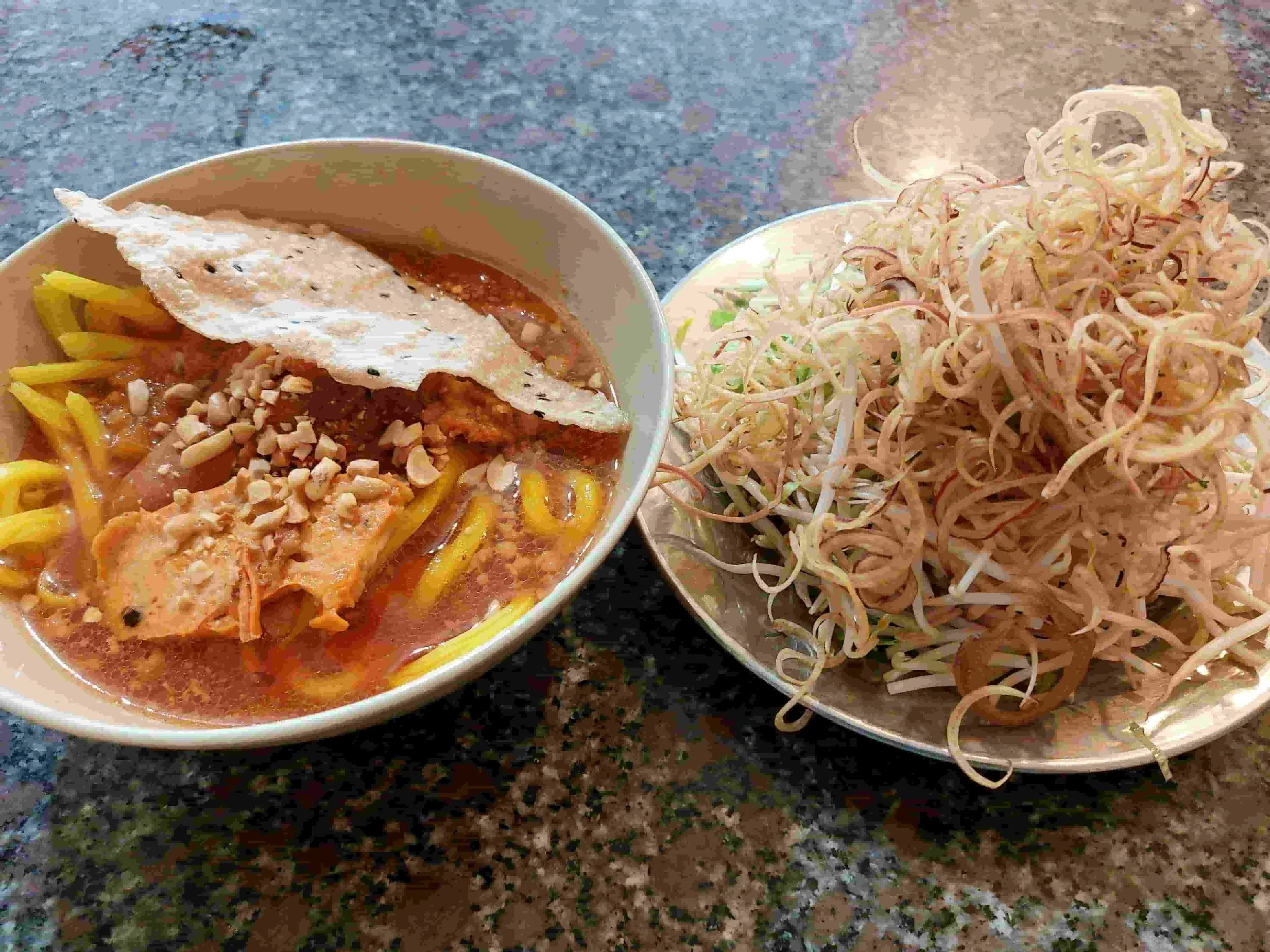 Mì Quảng – Vietnamese turmeric rice noodles with pork and shrimp, topped with crushed peanuts and crispy rice cracker, served with fresh herbs and vegetable sides