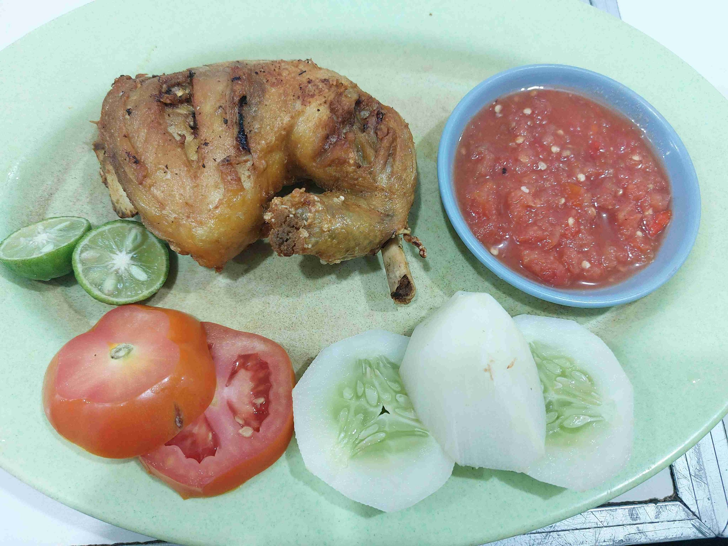 Indonesian ayam goreng fried chicken served with spicy sambal, fresh tomatoes, cucumber slices, and lime wedge
