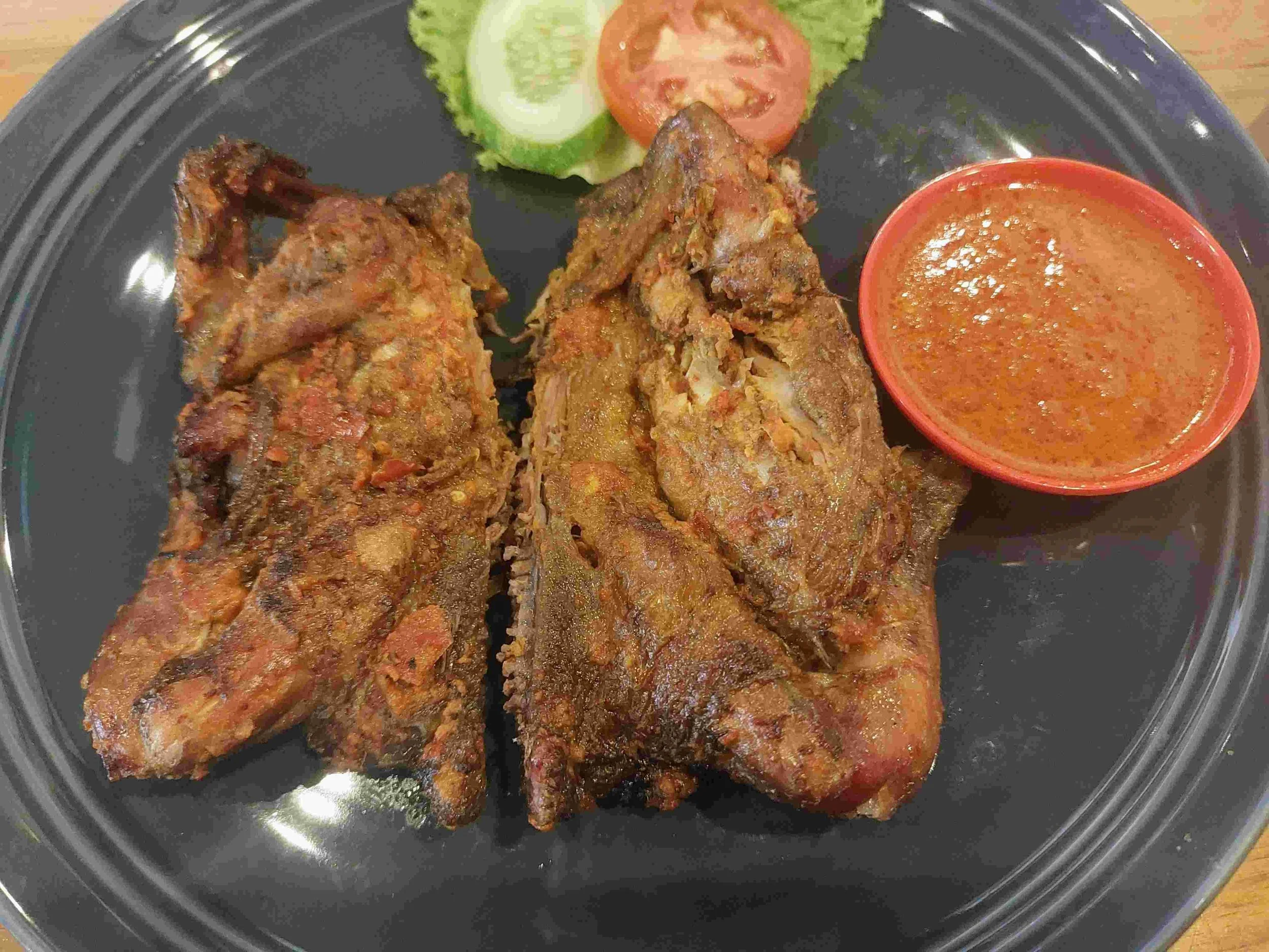 Ayam Taliwang grilled chicken coated in fiery chili sauce, accompanied by side sambal, cucumber, and tomatoes