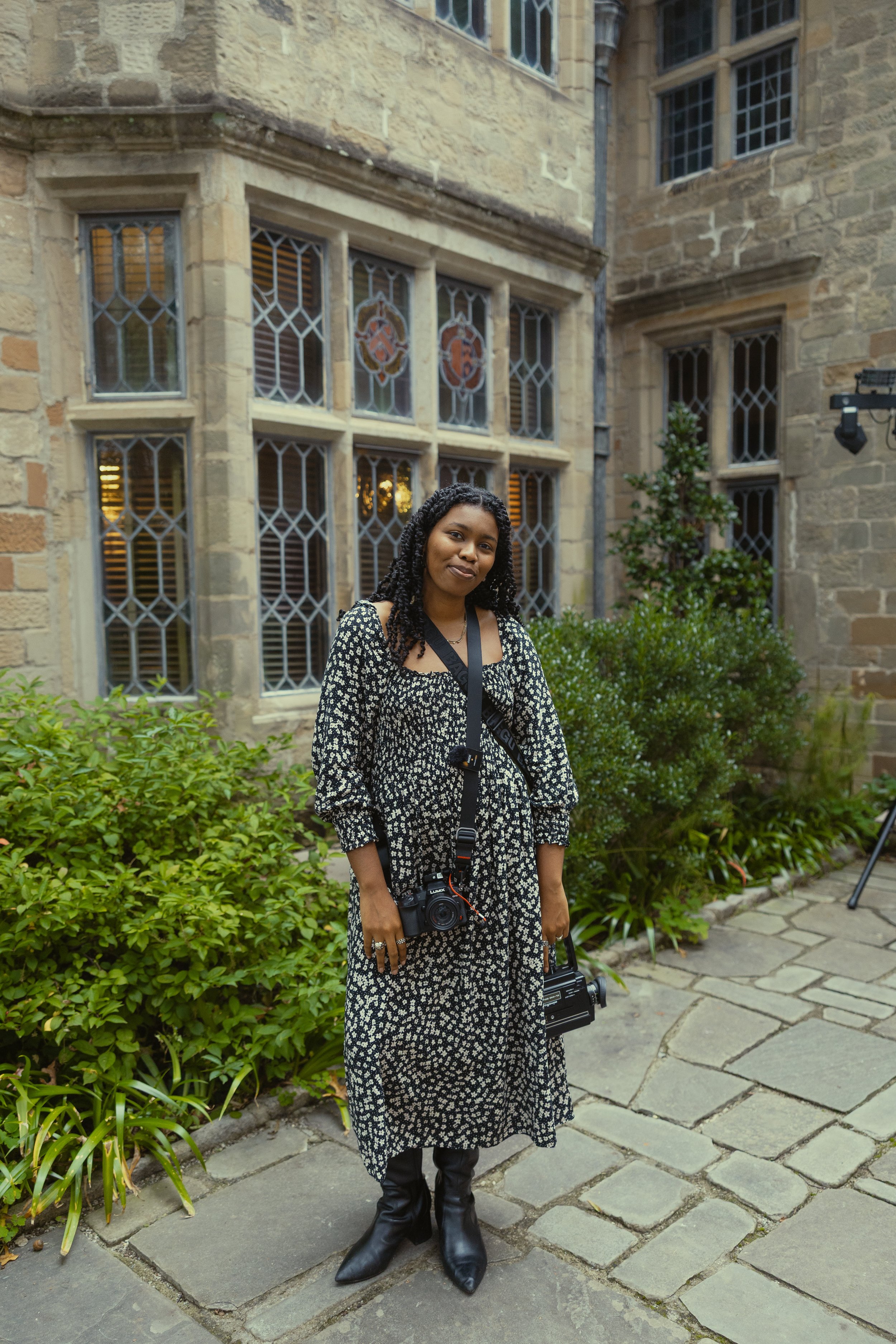 A woman with black curly hair wearing a black and white floral dress and black boots, standing outdoors on a stone-paved pathway in front of a historic stone building with stained-glass windows, greenery, and a tripod on the side.