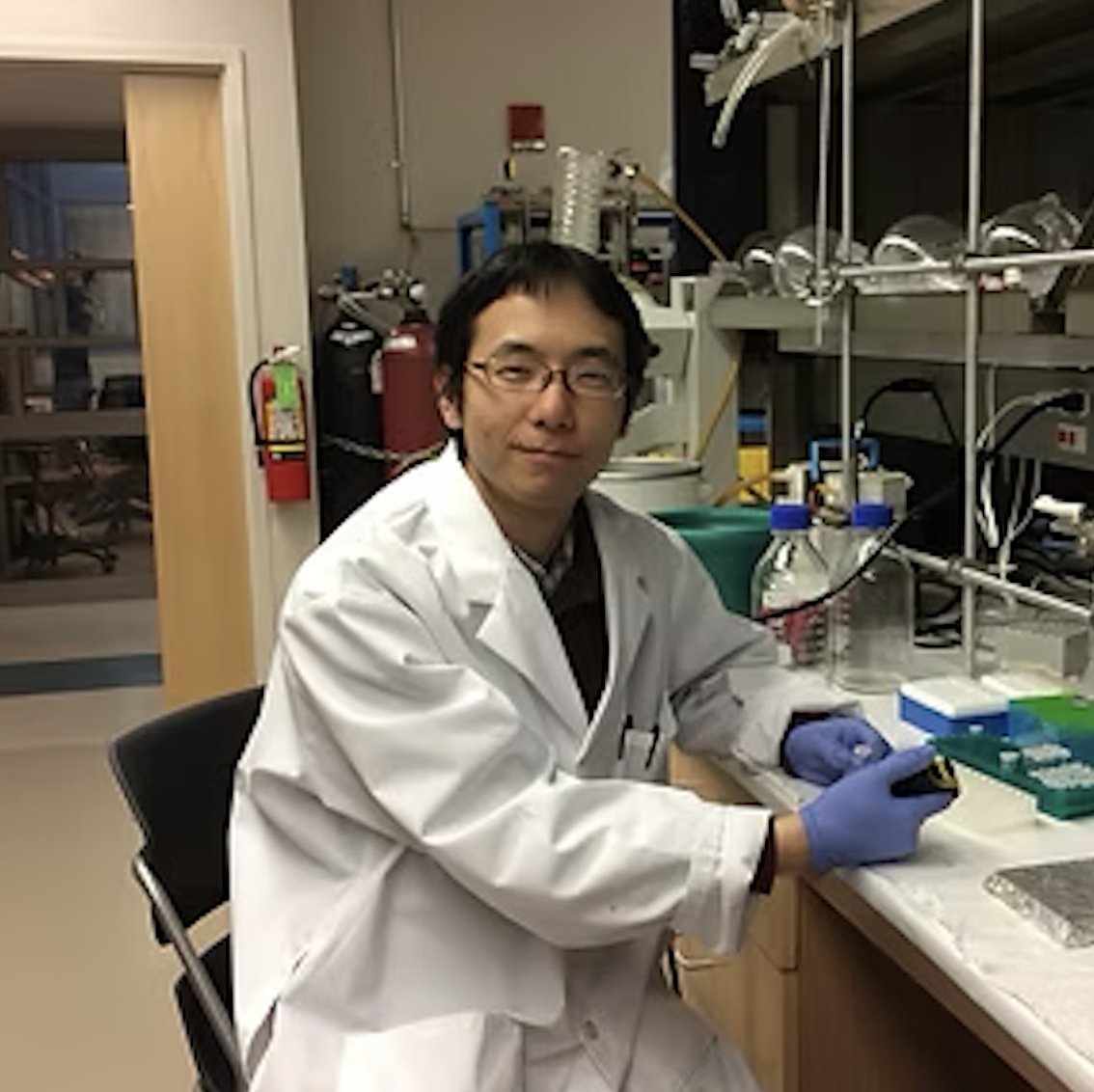 A scientist in a laboratory wearing a white lab coat, gloves, and glasses, sitting at a workbench with scientific equipment, bottles, and containers.