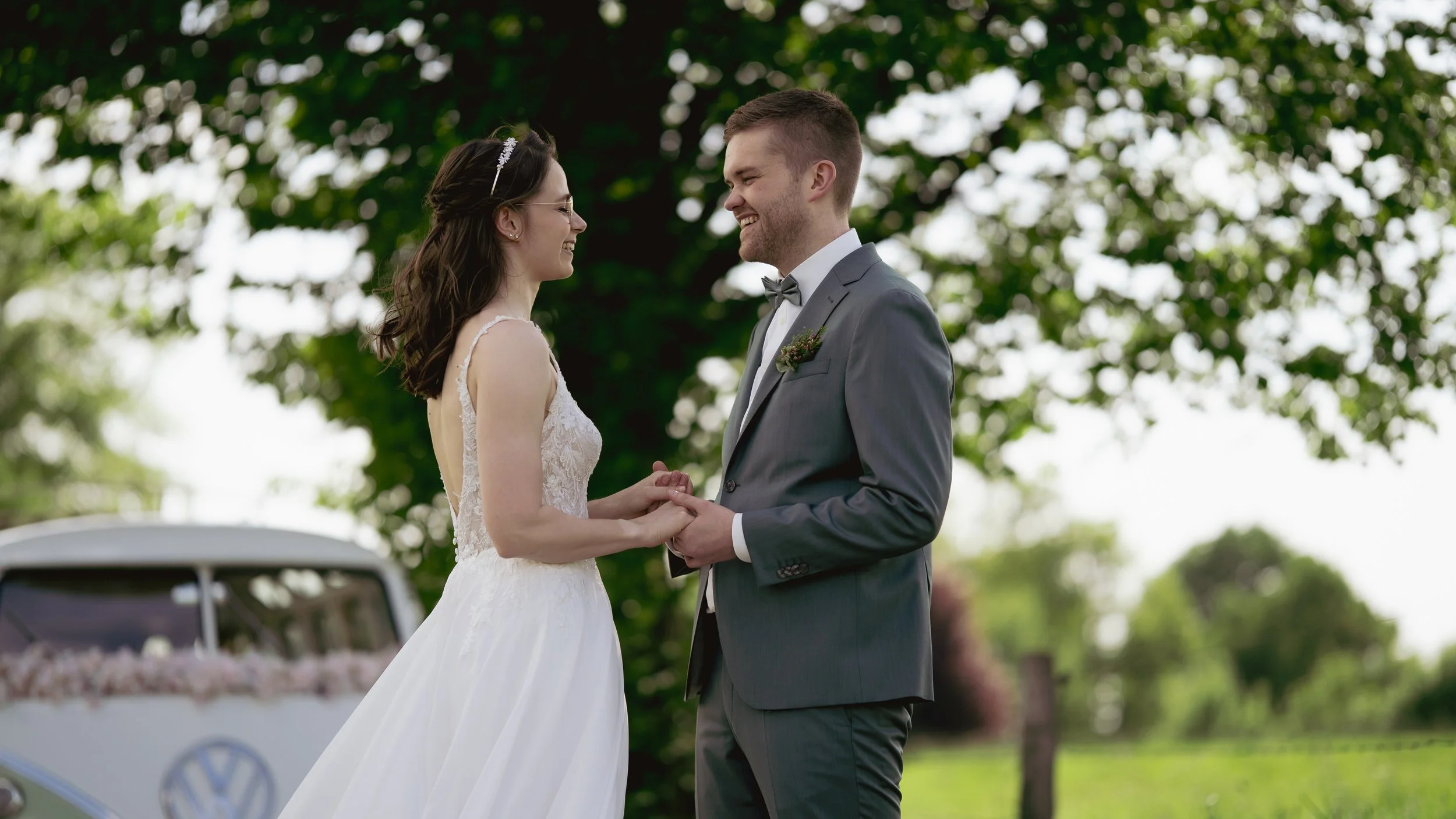 Ein Brautpaar steht sich bei einer Hochzeit im Freien gegenüber, lächelnd und Händchen haltend, vor grünen Bäumen, bei sonnigem Wetter.