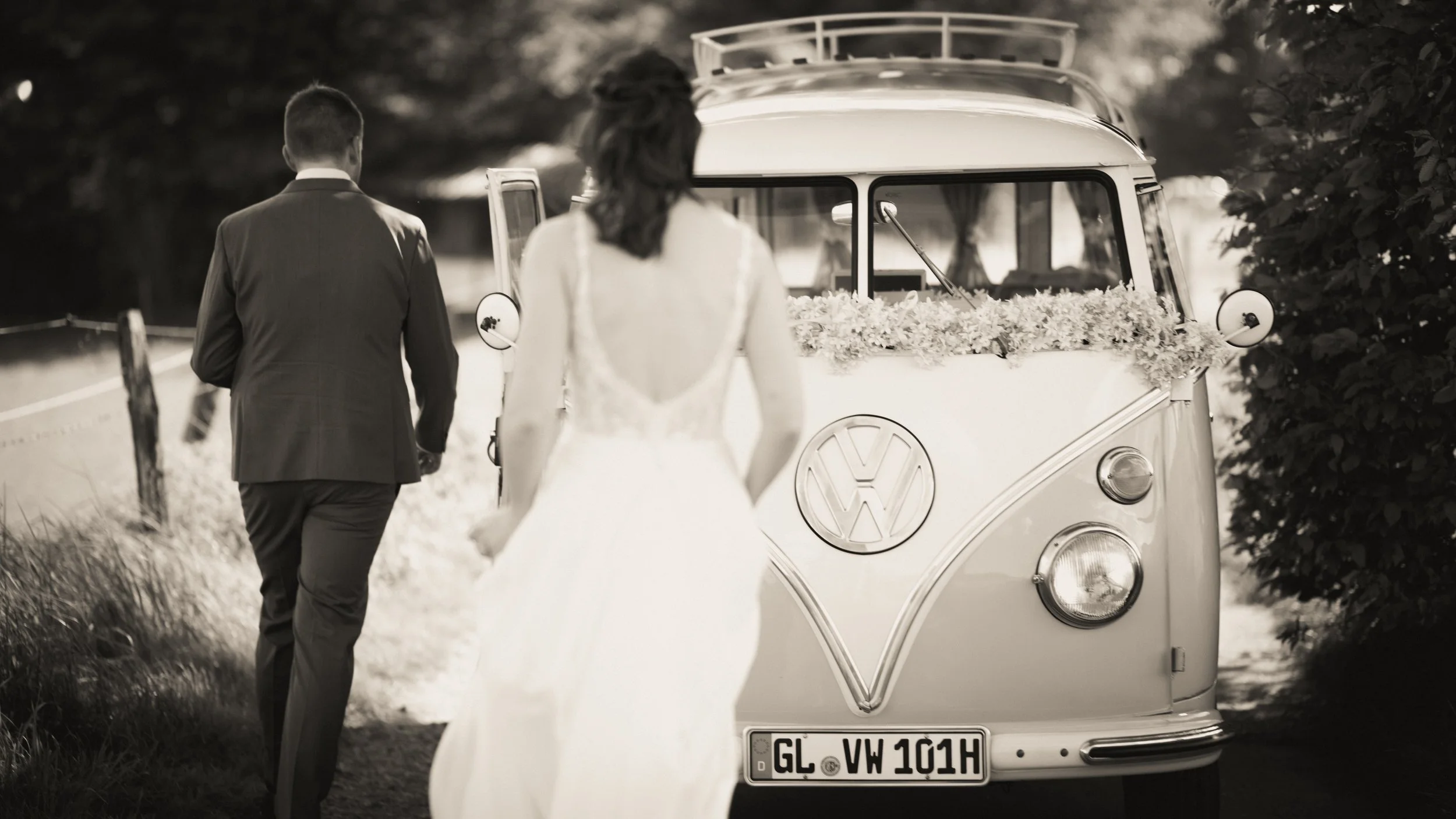 Ein Brautpaar geht an einem vintage VW-Campingbus vorbei, der mit Blumen dekoriert ist, in einer natürlichen Outdoor-Umgebung.