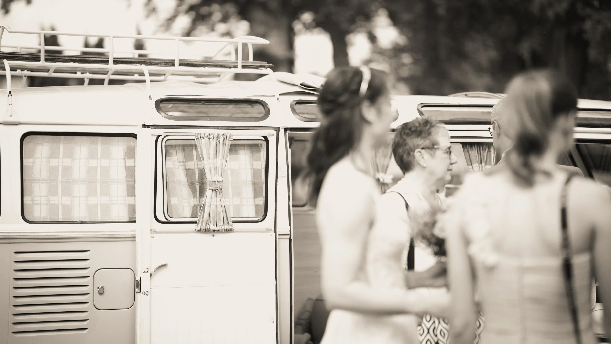 Schwarz-weiß-Foto von mehreren Menschen bei einer Veranstaltung im Freien, im Hintergrund ein Vintage-Camper mit geöffneten Fenstern und einem Vorhang.