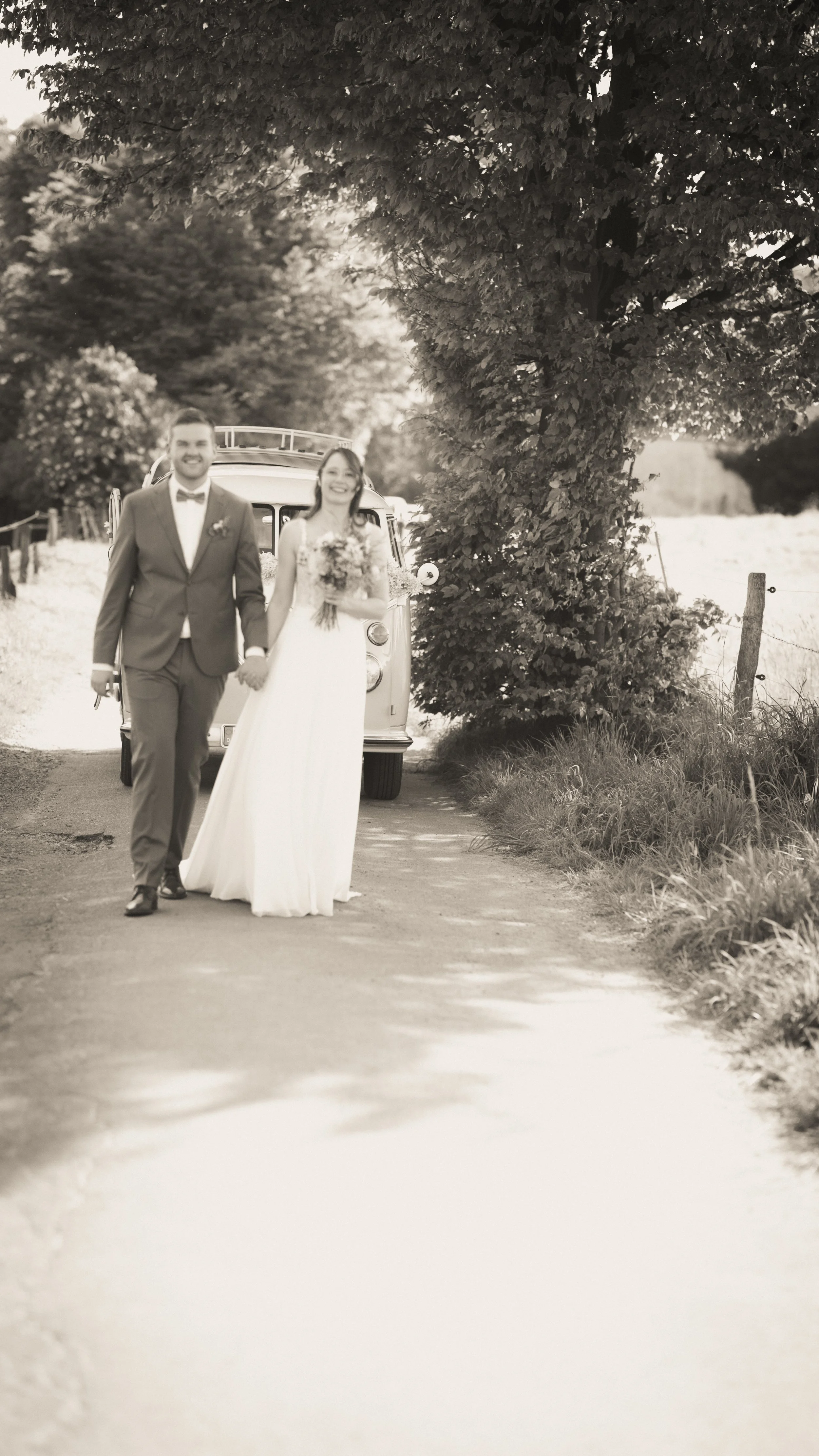 Ein Brautpaar, ein Mann in Anzug und eine Frau in Brautkleid, läuft Hand in Hand auf einem Landweg, im Hintergrund ein Oldtimer. Die Szene ist in Schwarzweiß gehalten.