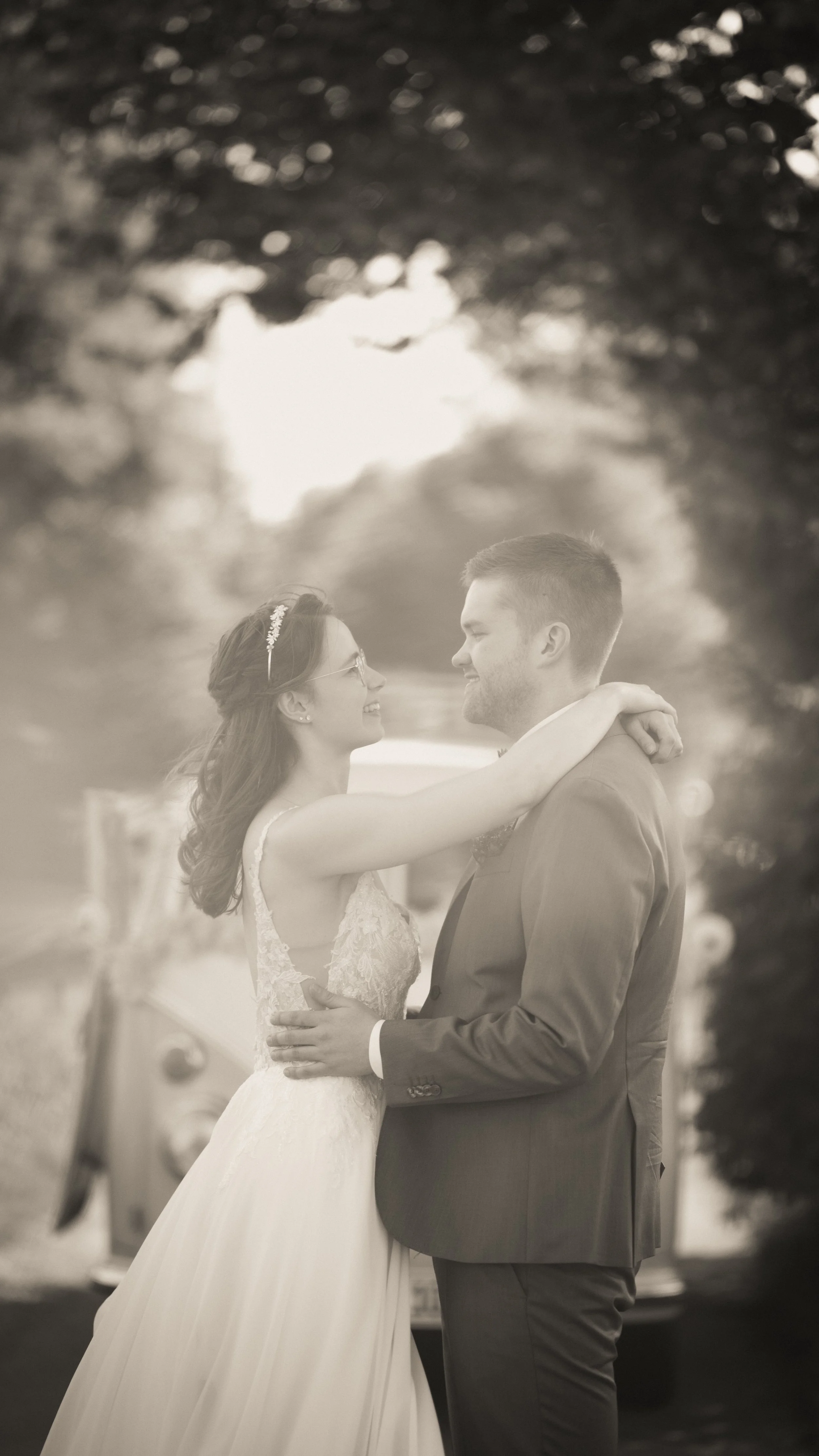 Ein Brautpaar bei einer Hochzeit, die Frau trägt ein weißes Brautkleid mit Spitzendetails und eine kleine Tiara, der Mann trägt einen Anzug. Sie schauen sich liebevoll an, während sie sich umarmen.