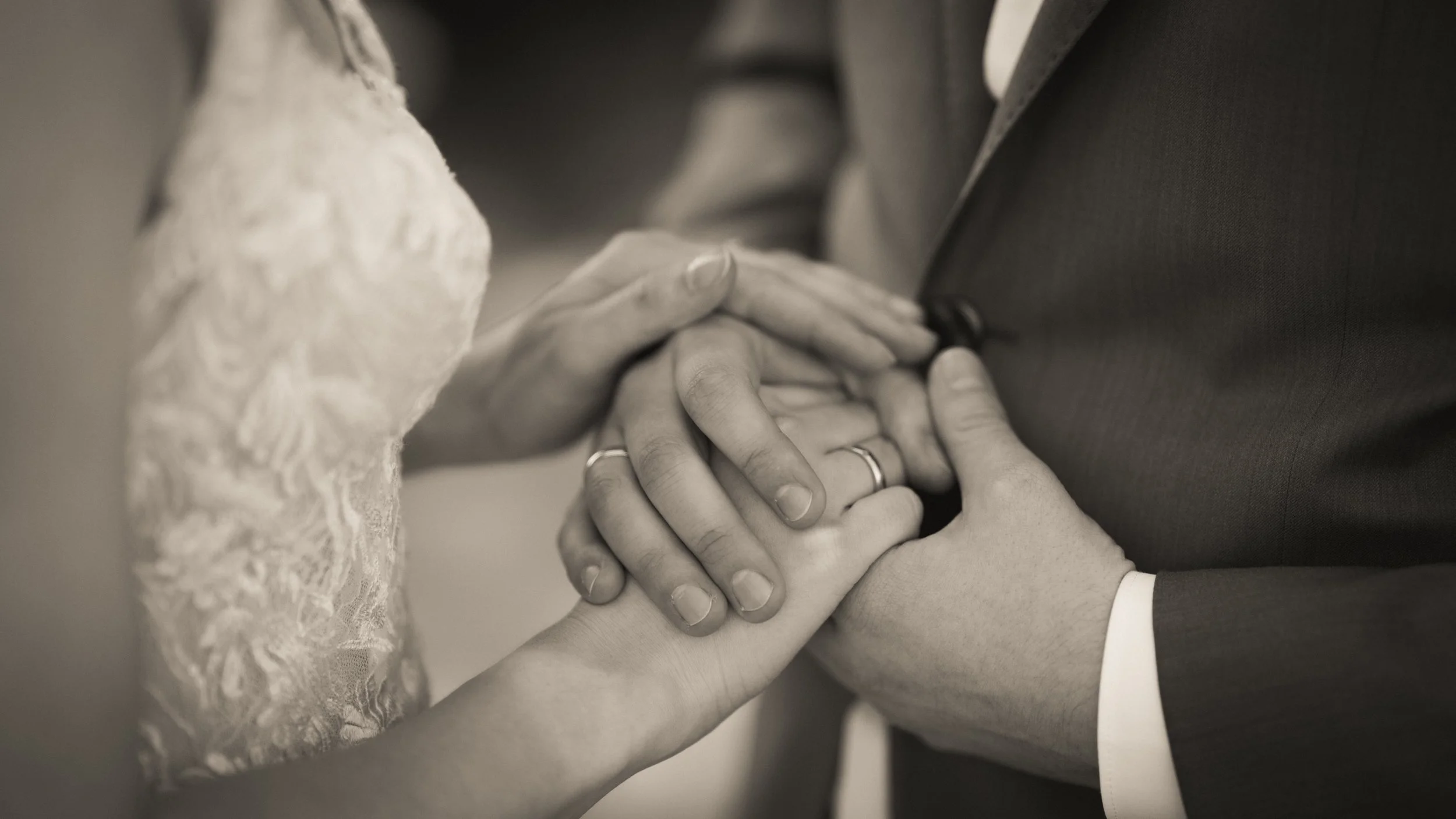 Nahaufnahme von Händen, die sich auf einer Hochzeit halten, mit einem Brautkleid und Anzug im Hintergrund.
