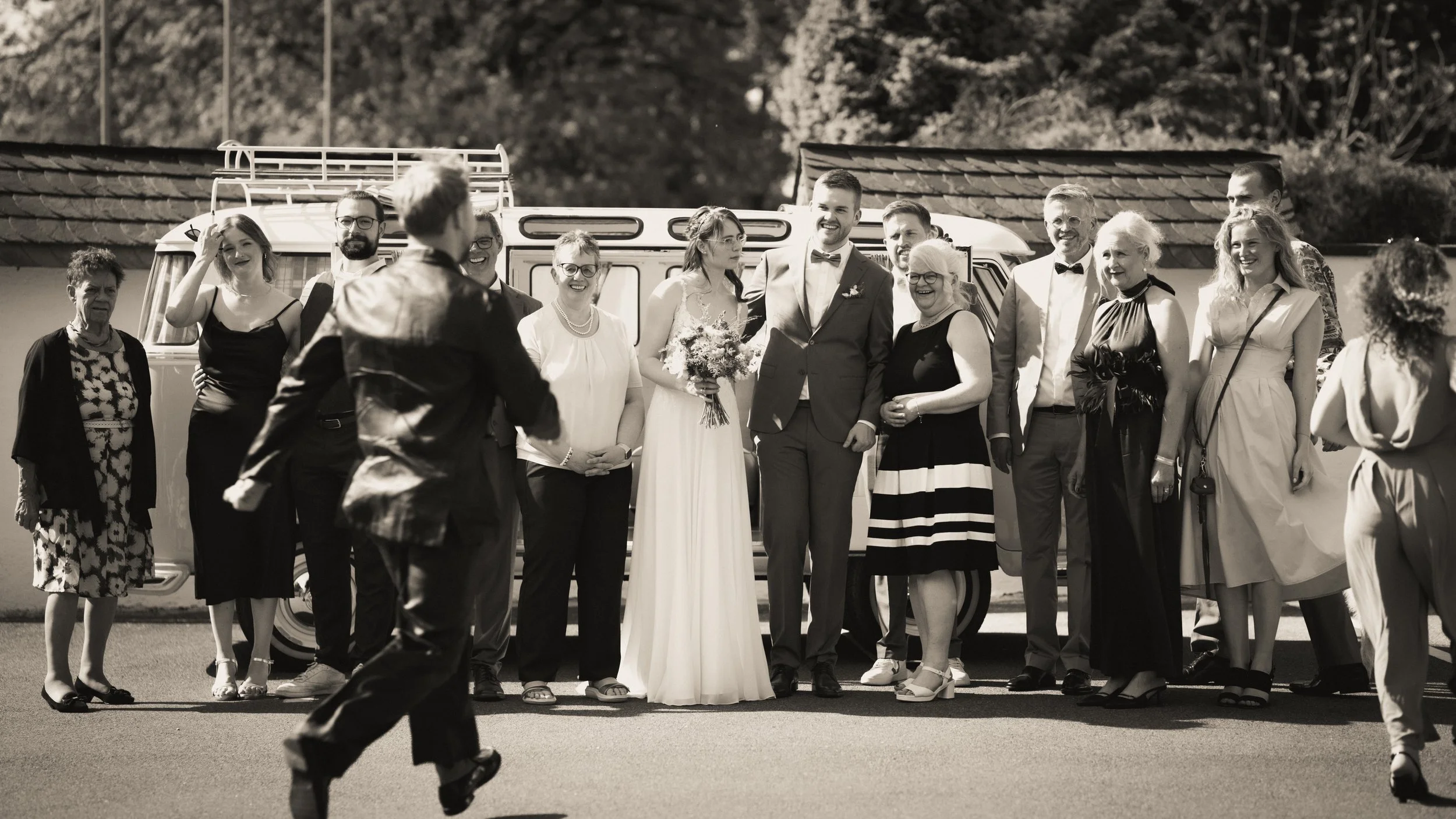 Eine Hochzeitsfeier mit der Hochzeitspaar im Mittelpunkt, umgeben von Gästen, vor einem Wohnmobil, schwarz-weiße Fotografie.