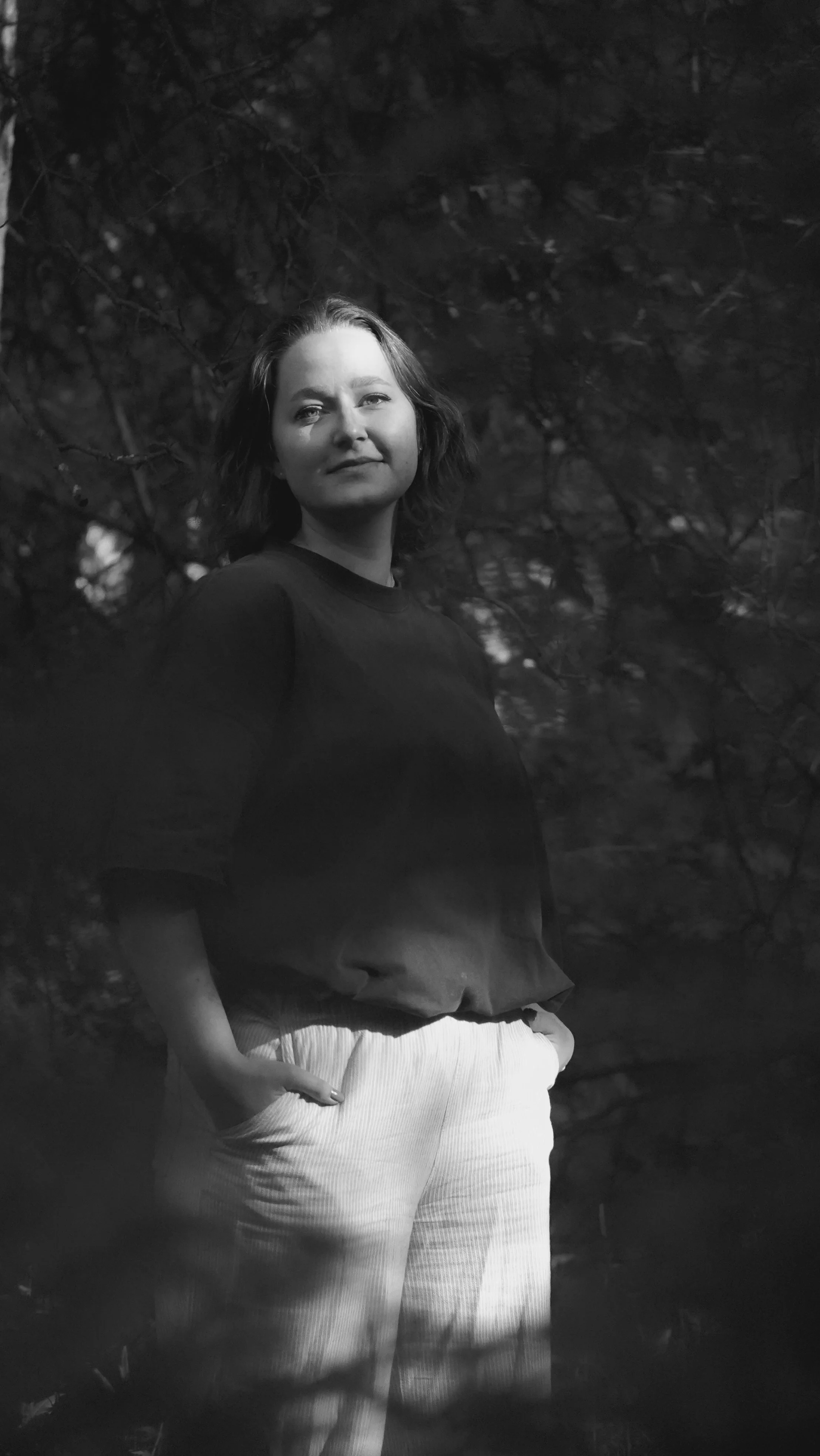Schwarz-weiß-Foto einer jungen Frau mit kurzen Haaren, die in die Kamera blickt und an einem dunklen Ort mit Bäumen im Hintergrund steht, ihre Hände in den Taschen ihrer weißen Hose.