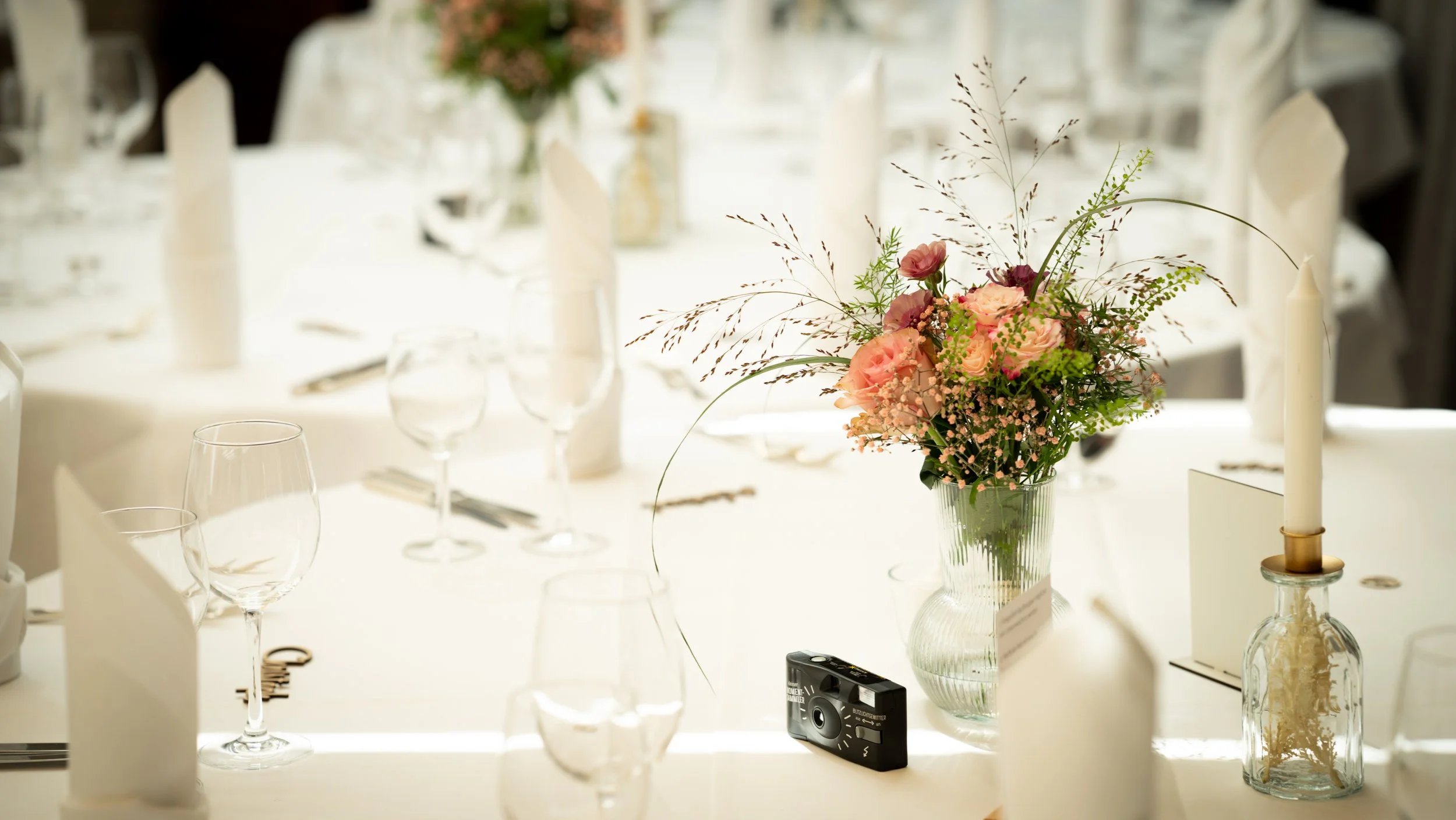 Blumenstrauß mit rosa und lila Blumen auf einem gedeckten Tisch, umgeben von Gläsern und Kerzen, bei einer eleganten Veranstaltung.