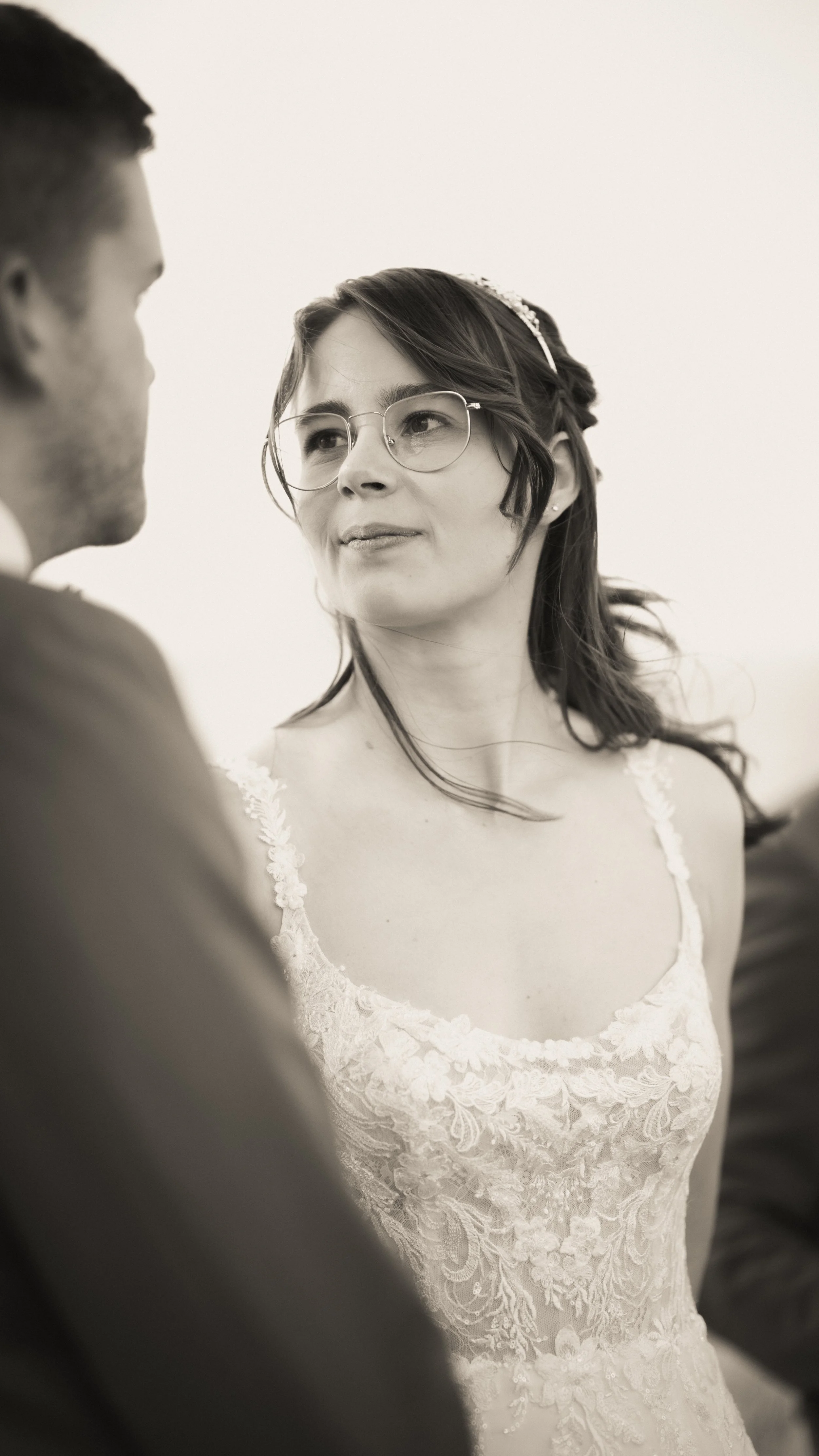 Eine Braut im weißen Kleid mit Spitze schaut zu einem Mann, der nur teilweise sichtbar ist, auf einer Hochzeit.