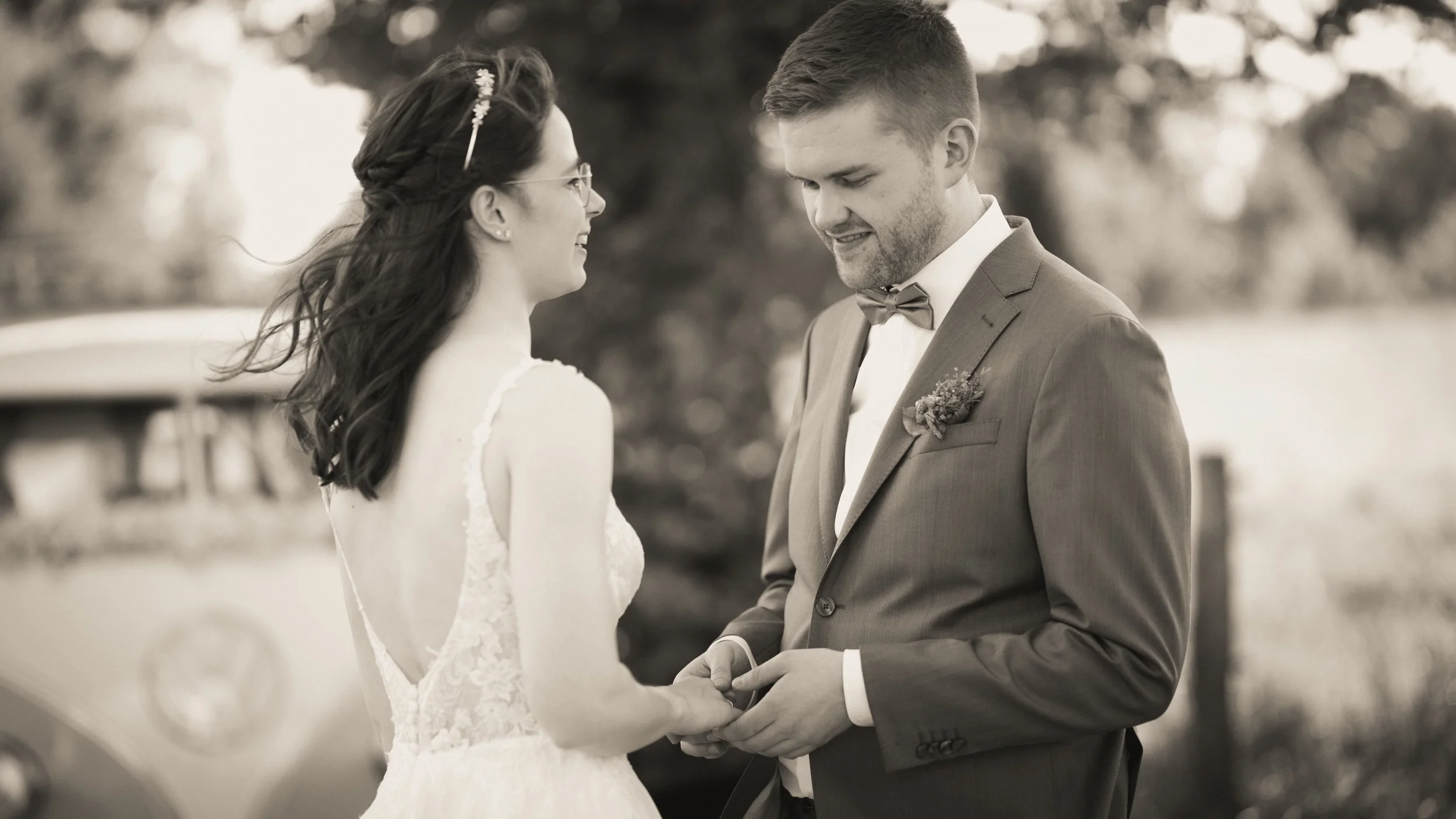 Das Bild zeigt ein Brautpaar bei ihrer Hochzeit, das sich liebevoll anblickt und Händchen hält. Das Brautkleid ist weiß und hat Lace-Details, der Bräutigam trägt einen Anzug mit Fliege. Es ist eine emotionale Szene im Freien.