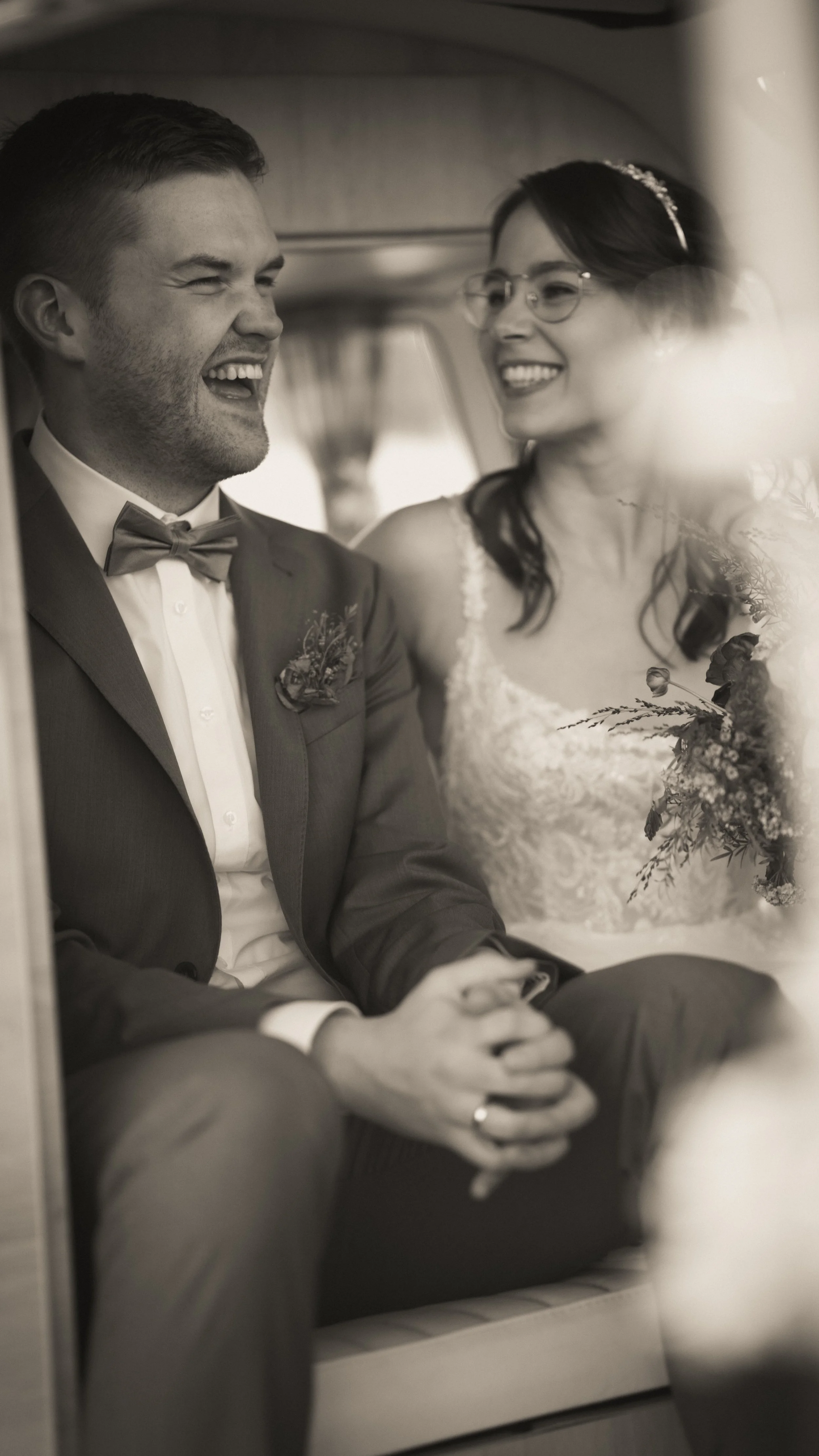 Ein Brautpaar sitzt in einem Fahrzeug, lacht und hält Händchen, Hochzeitskleidung, schwarz-weiße Aufnahme.