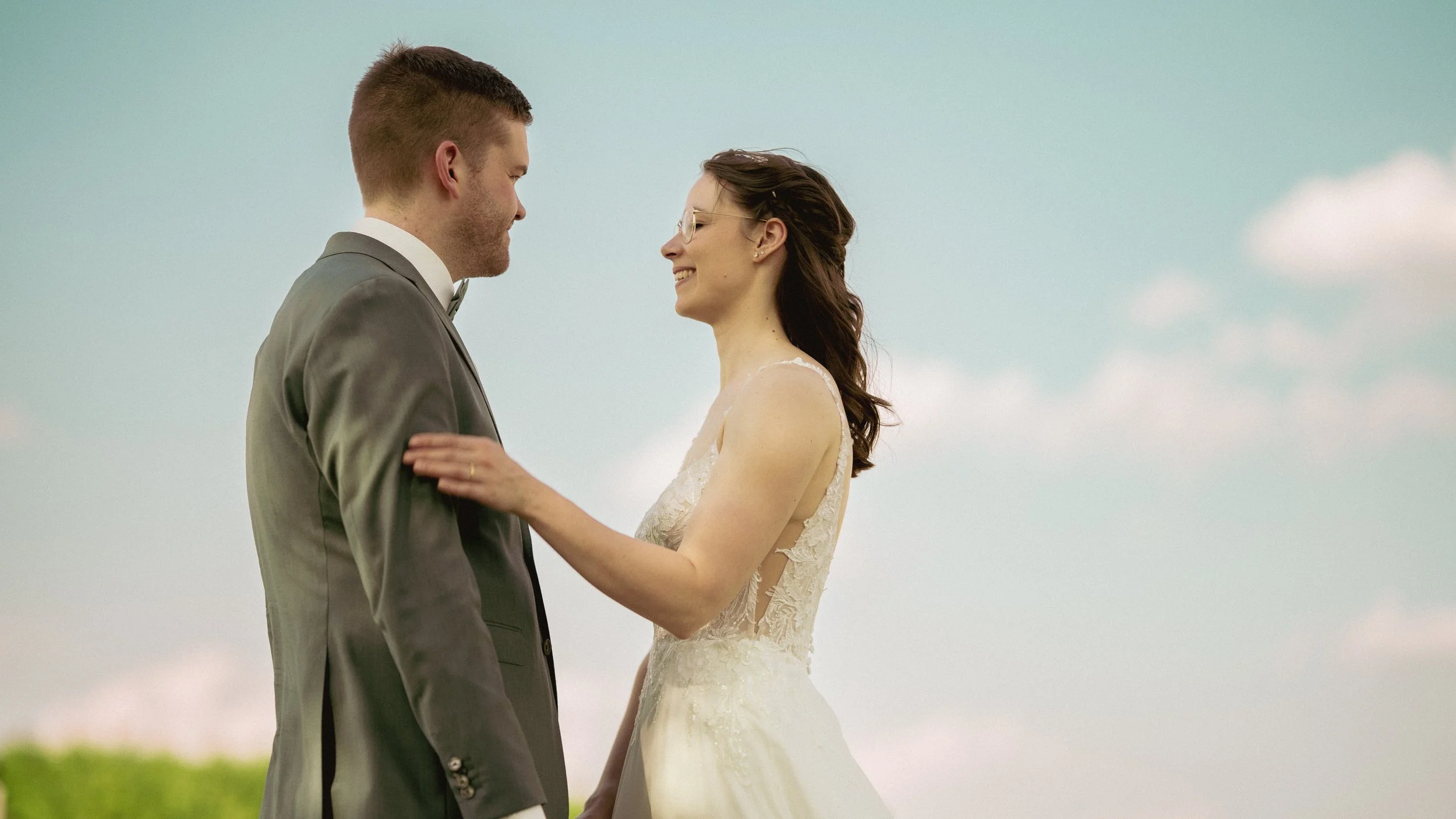 Ein Paar bei einer Hochzeit, das sich liebevoll ansieht und sich die Hände hält, im Freien mit Himmel im Hintergrund.