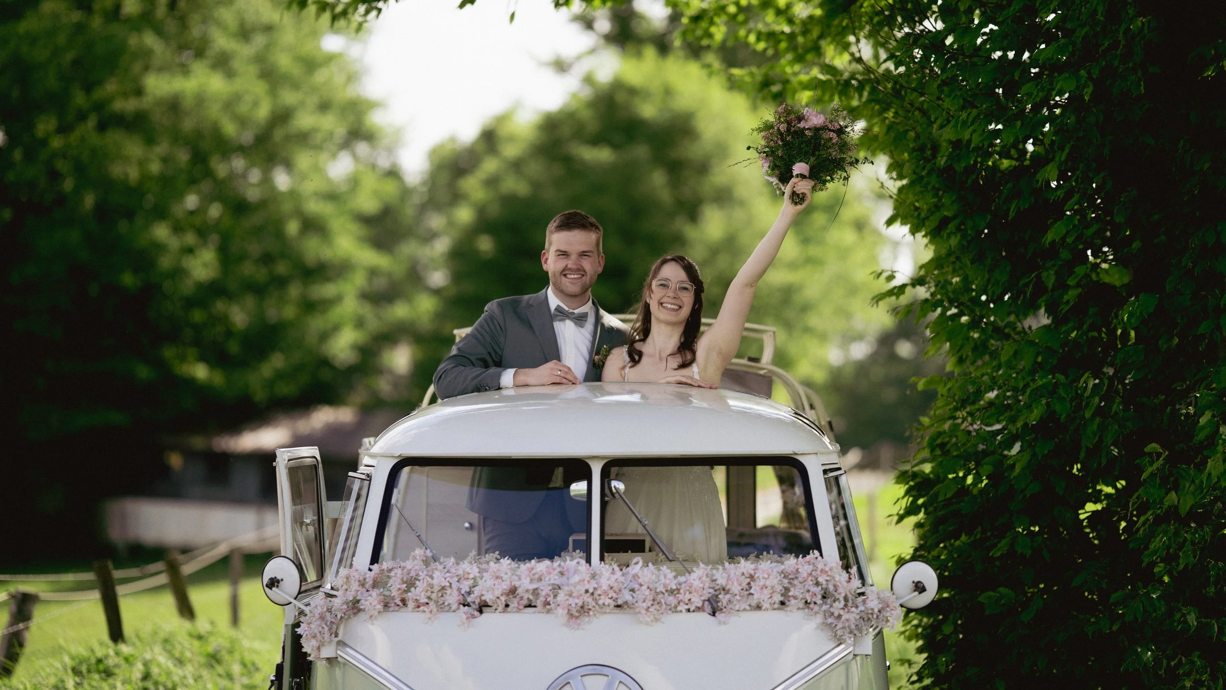 Ein frisch verheiratetes Paar fährt in einem weißen Oldtimer, der mit Blumen geschmückt ist, durch eine grüne Parklandschaft. Der Bräutigam trägt einen schwarzen Anzug mit Fliege, die Braut ein weißes Kleid, sie hält einen Blumenstrauß hoch und läche