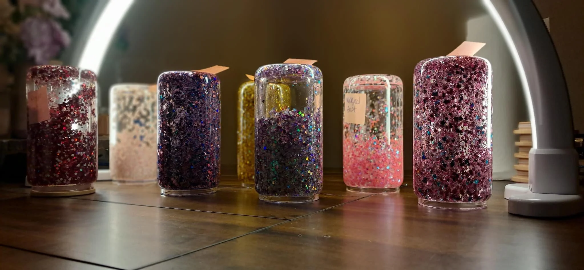 Several cylindrical glitter-filled resin tumblers with various colors and glitter patterns placed on a wooden table under a circular light.