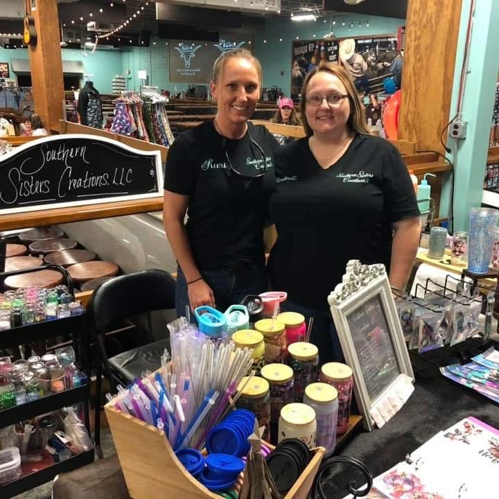 Two women standing behind a table filled with jars, pens, and craft supplies inside a store called Southern Sisters Creations LLC.
