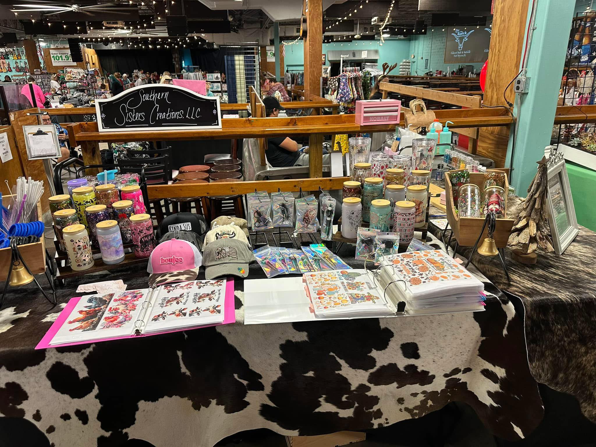 A craft vendor table at an indoor market displays various stickers, hats, candles, and stationery with a sign reading "Southern Sisters Creations LLC." in the background. The market has wooden railings, string lights, and multiple shoppers.