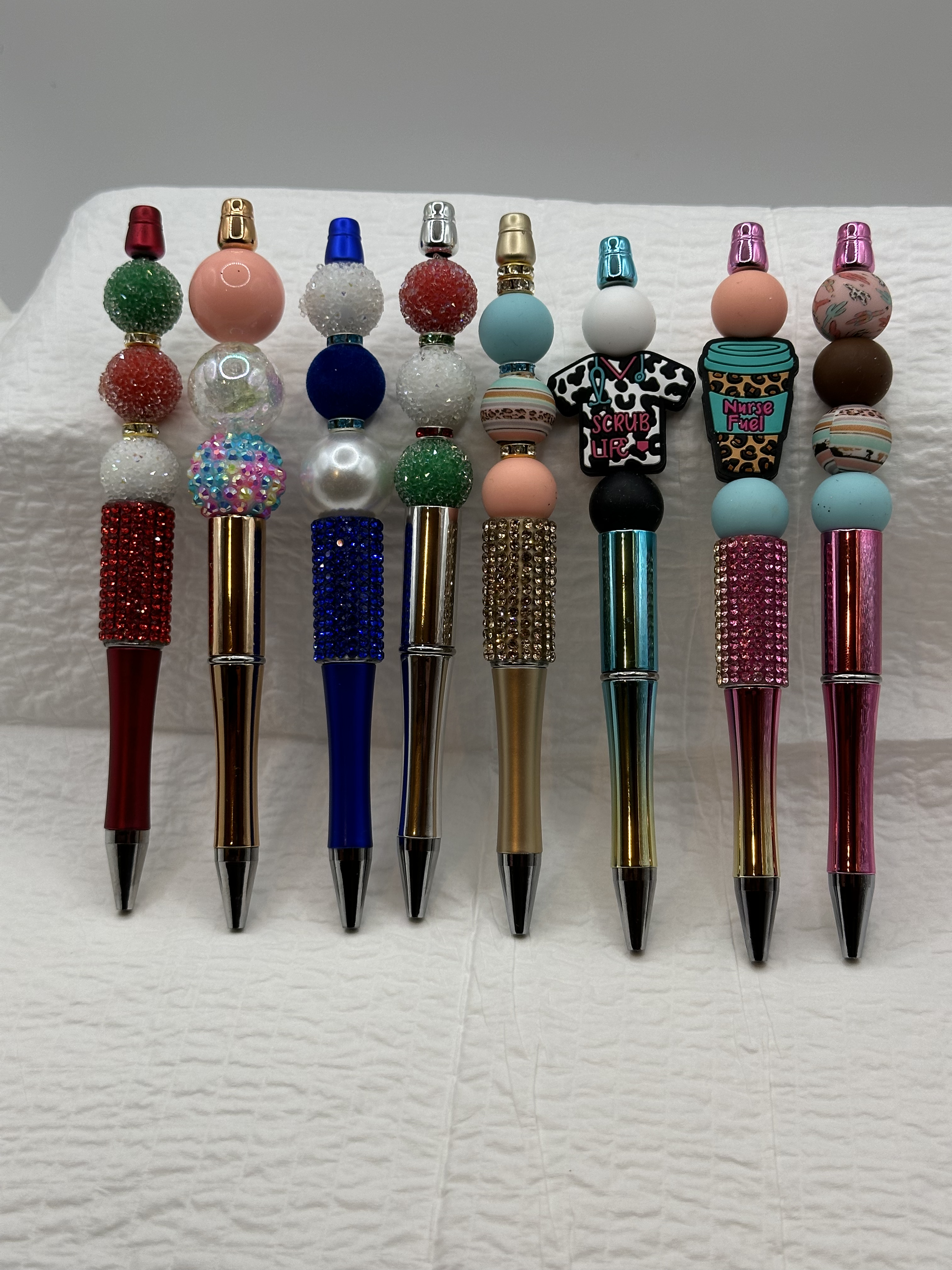 Collection of pens with decorative tops featuring various colors, textures, and themed charms, including scrubs and nurse symbols.