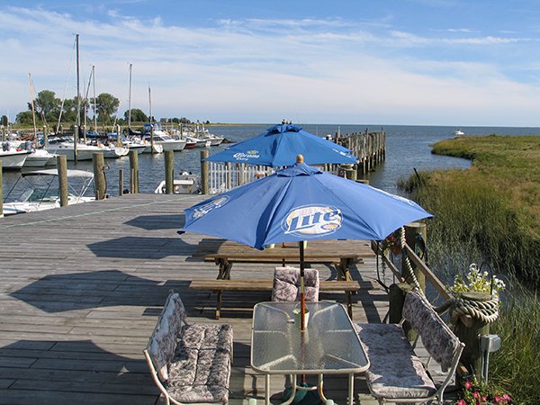 Dockside seating with tables and blue umbrellas, overlooking a marina with boats and water.