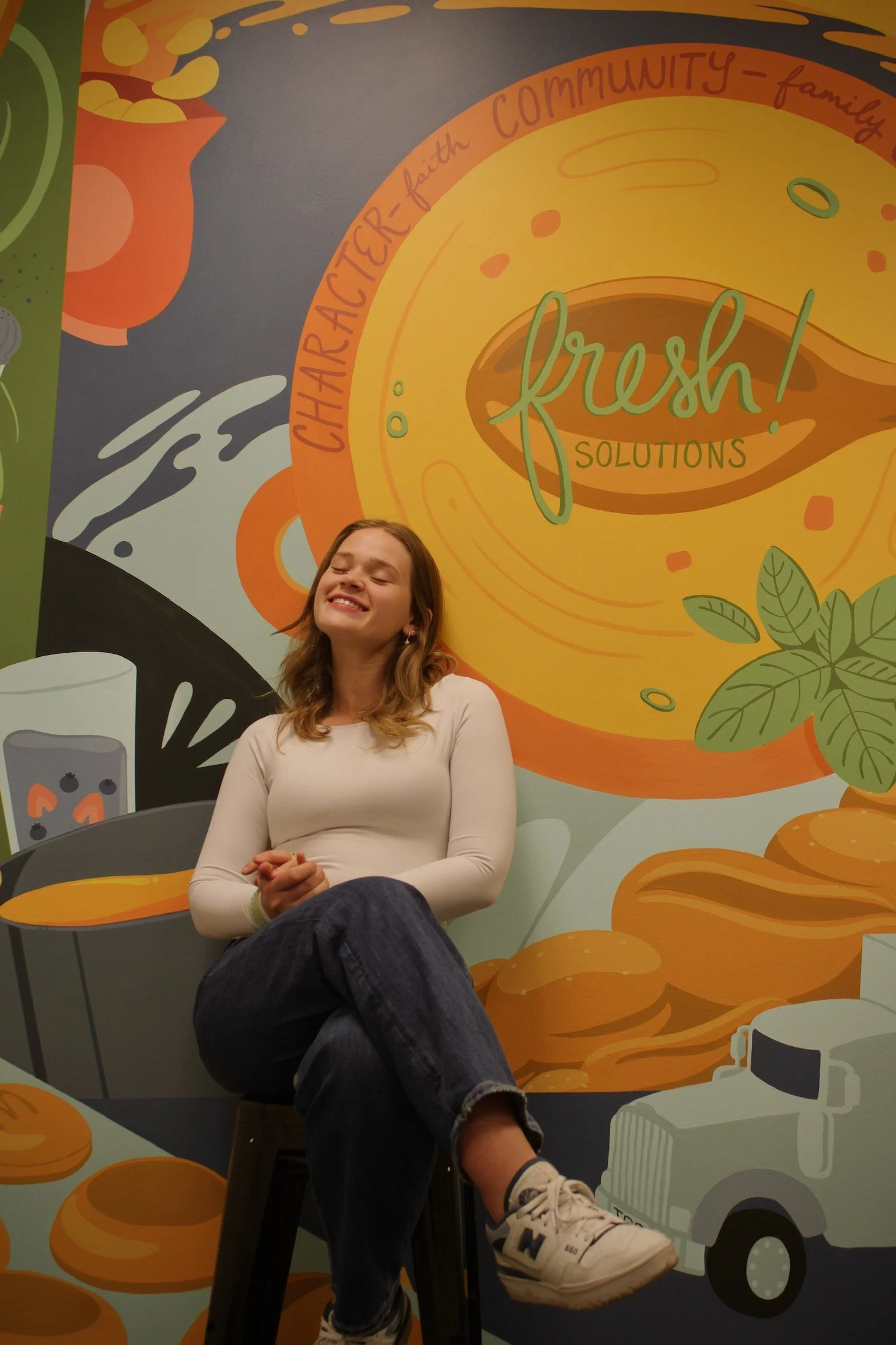 A young woman with long hair, wearing a white long-sleeve shirt and jeans, sitting on a black stool with her eyes closed and smiling, in front of a colorful mural with illustrations of food, a coffee cup, a delivery truck, and text that reads 'Fresh! Solutions' and other phrases related to community and family.