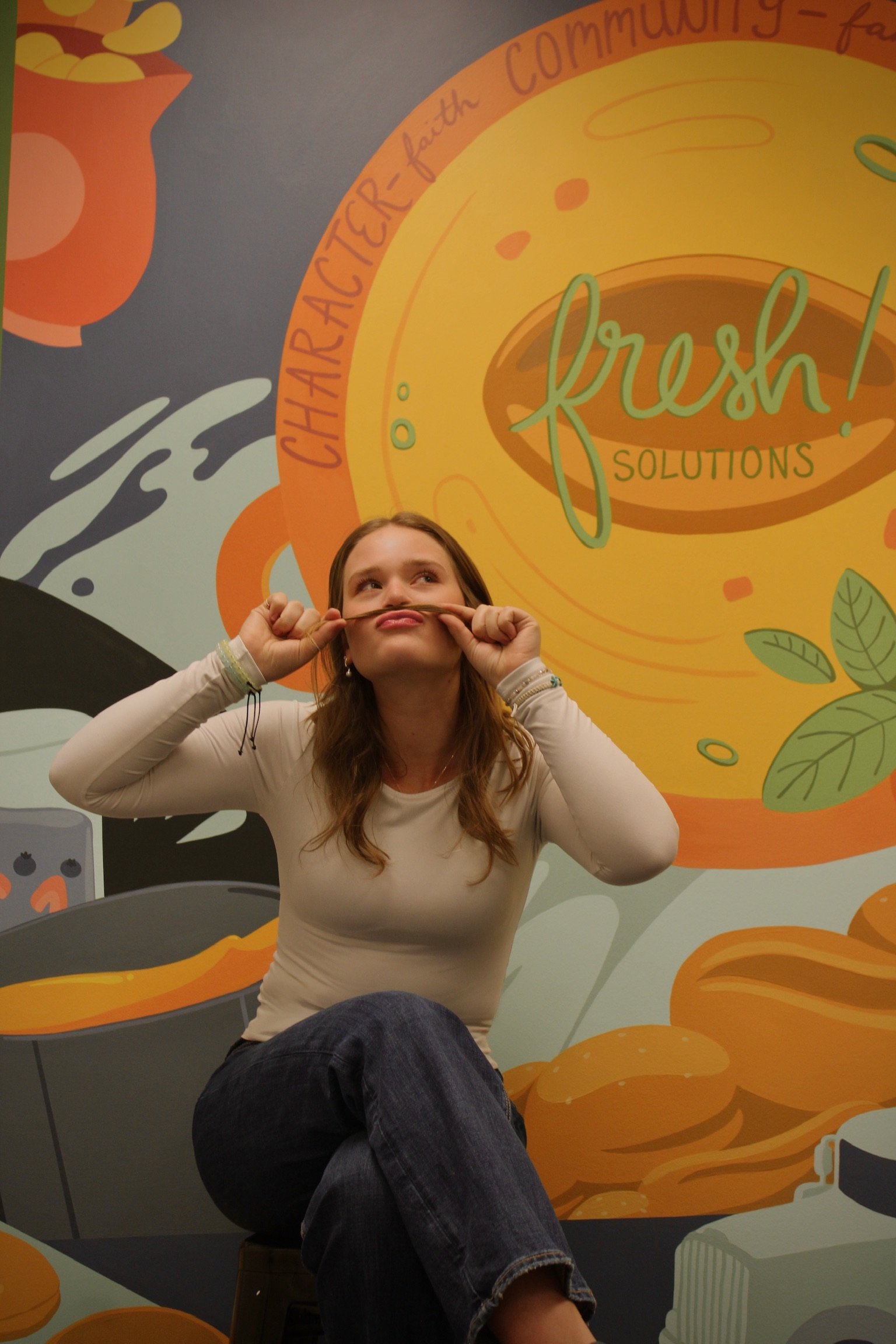 A young woman with long brown hair sitting and holding her glasses with a playful expression, in front of a colorful mural with the words 'fresh solutions' and related phrases like 'charactier-faith community' and illustrated food items.
