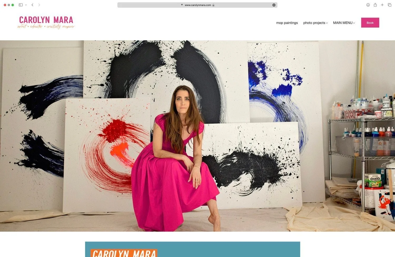 A woman in a bright pink dress sitting on the floor surrounded by abstract paintings with bold brushstrokes and splashes of black, red, and blue, in an artist's studio with paint bottles and supplies on a metal shelf to the right.