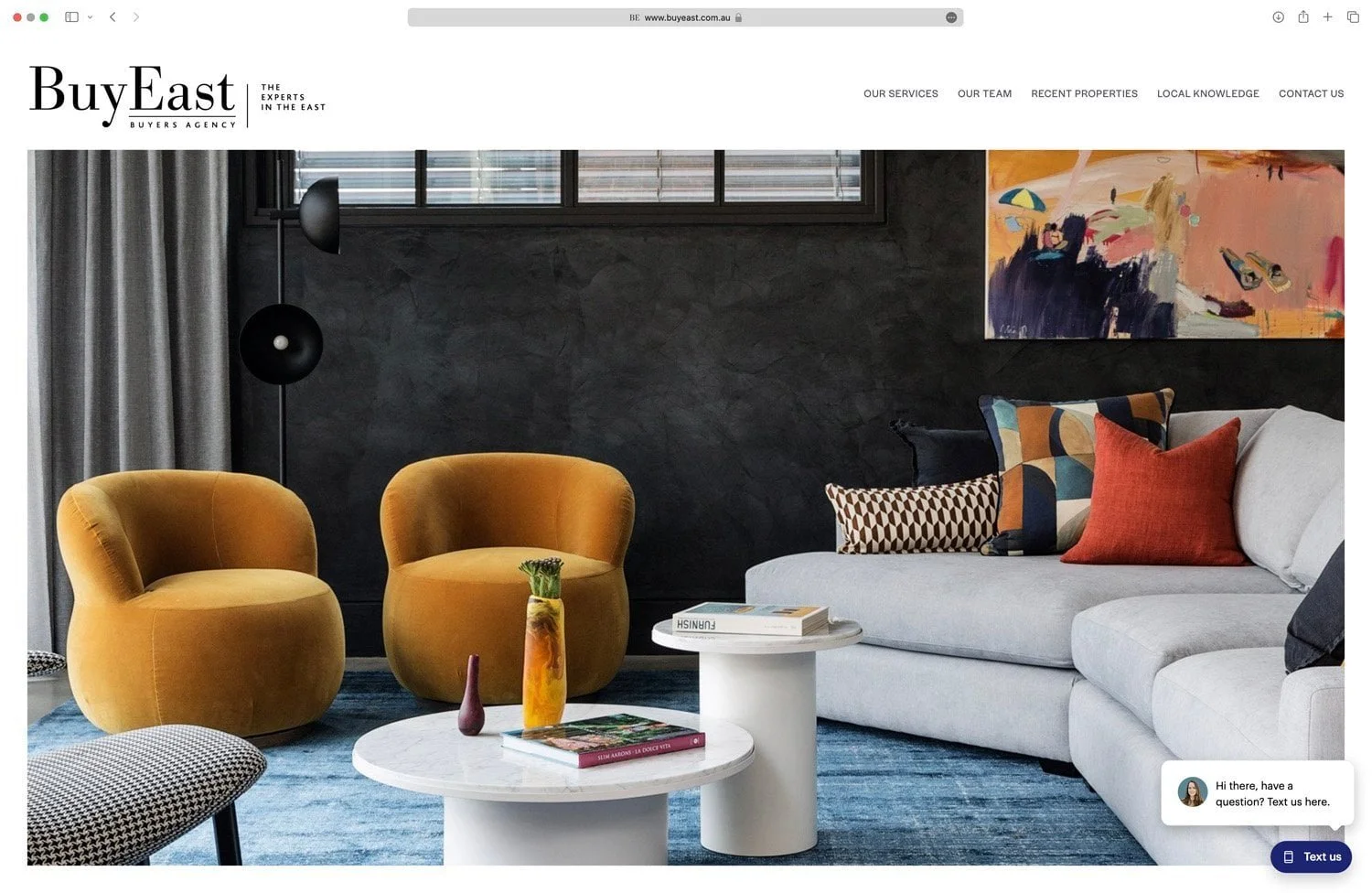 Living room with two yellow armchairs, a white sectional sofa with colorful cushions, a white round coffee table with decorative vases and books, a blue rug, a dark textured wall, a large window with blinds, a colorful abstract painting, and a floor lamp.