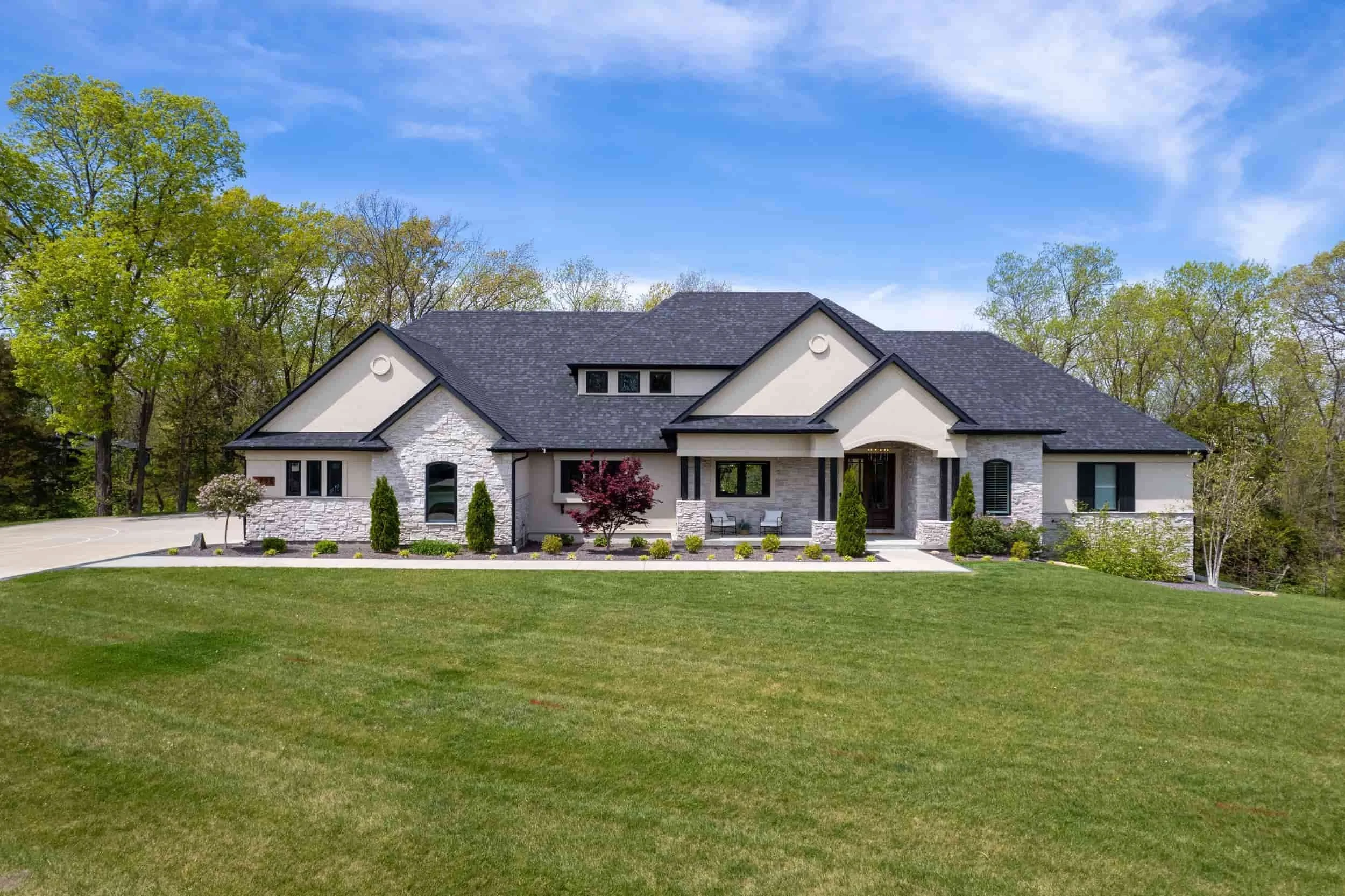 A large, modern house with a dark shingled roof and white stone and stucco exterior, surrounded by a well-maintained lawn and trees, under a partly cloudy sky.