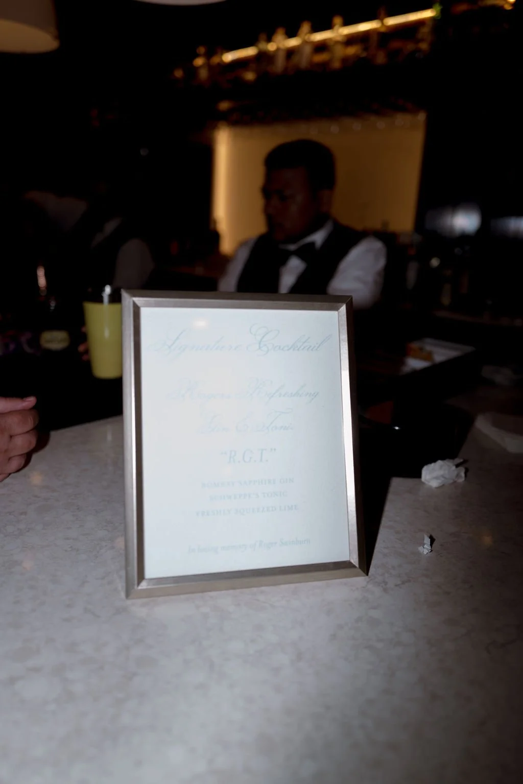 Framed menu on a bar counter with a bartender in the background, dimly lit setting.