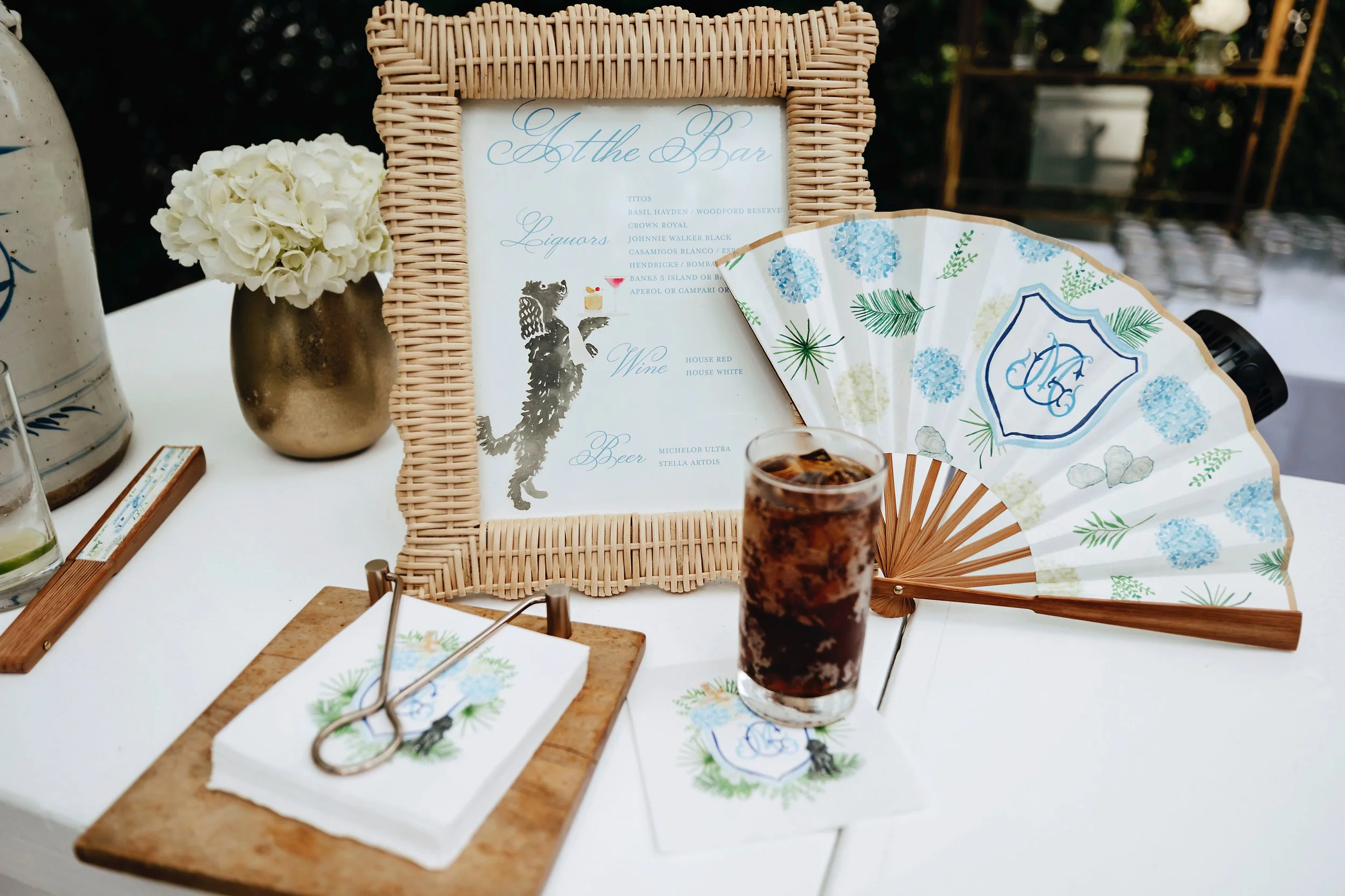 A cocktail menu framed in wicker, a paper fan with blue floral patterns, a glass of cola with ice, a gold vase with white flowers, and cocktail napkins with a monogram and tropical design on a white table.