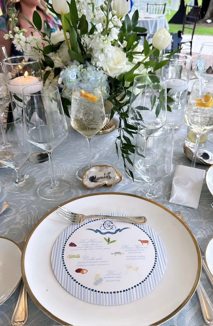 Elegant table setting with white plates, gold trim, silverware, and a floral centerpiece of white flowers and greenery. Multiple wine glasses with water and lemon slices, a candle, and a decorative name tag reading 'Madeline'. A printed menu on the p