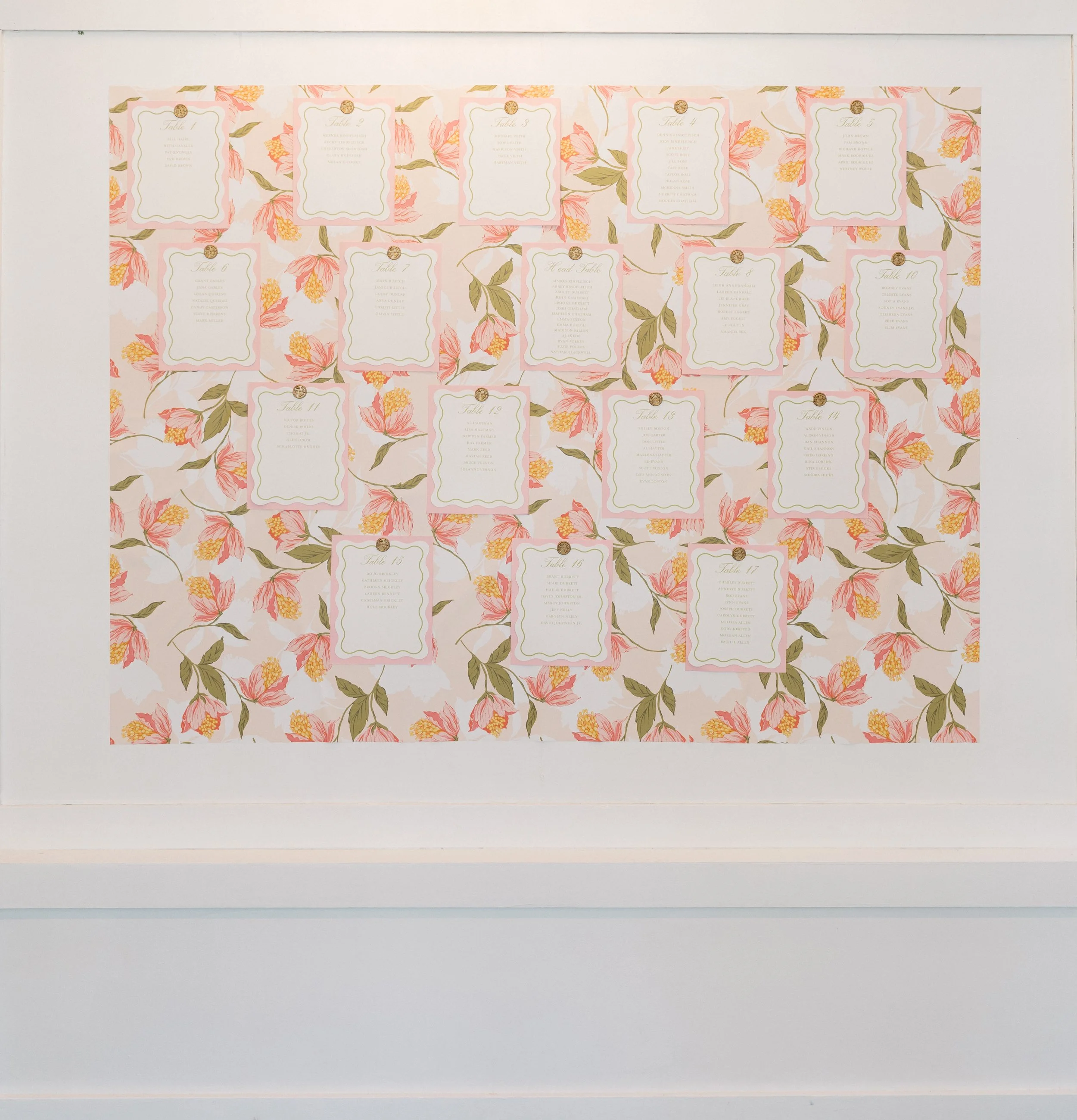 A wedding seating chart on floral patterned wallpaper background featuring numbered tables and guest names.