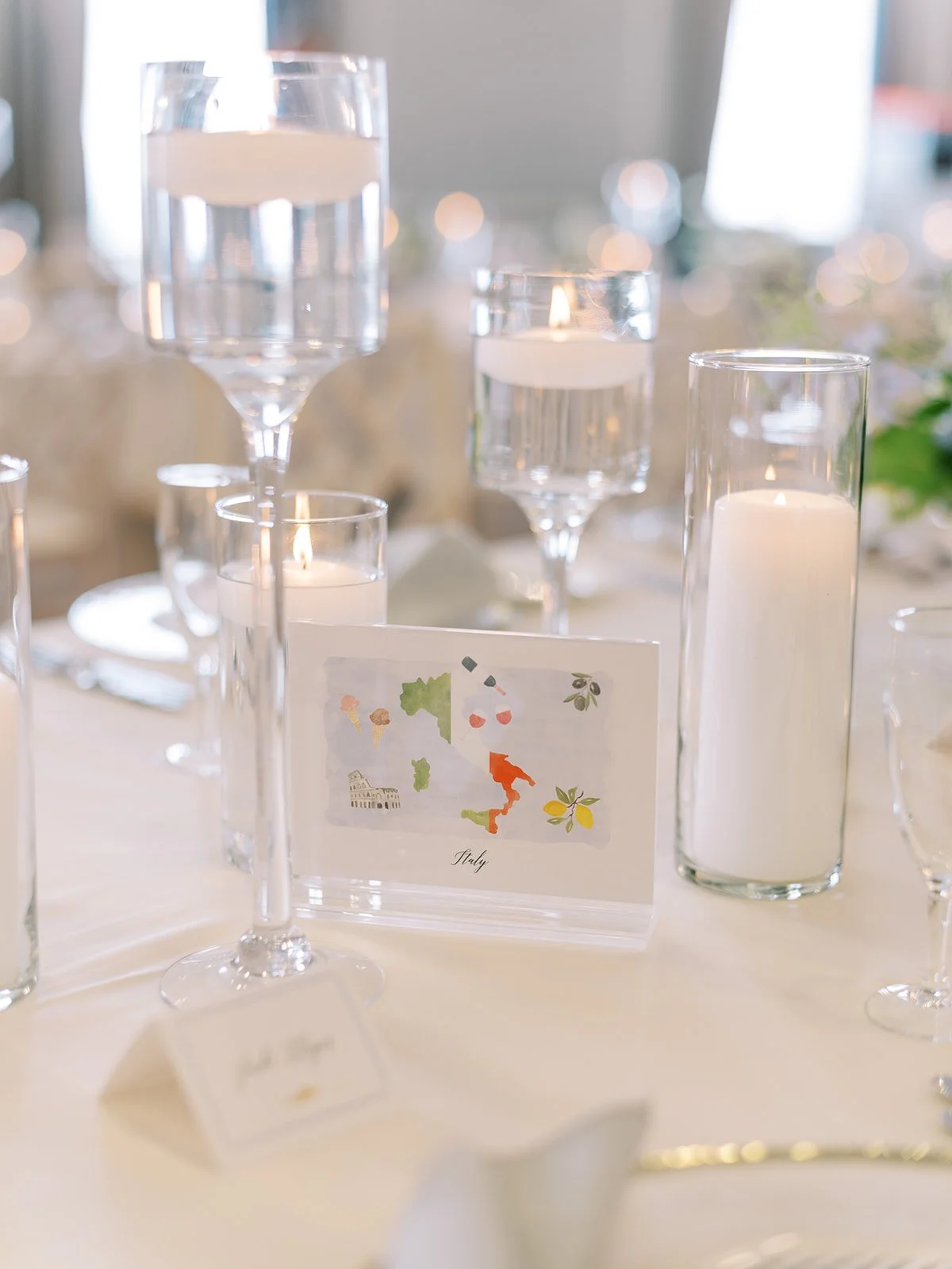 Table centerpiece with several glass candle holders containing white candles, a table card with a colorful map of Italy, and blurred background lights.