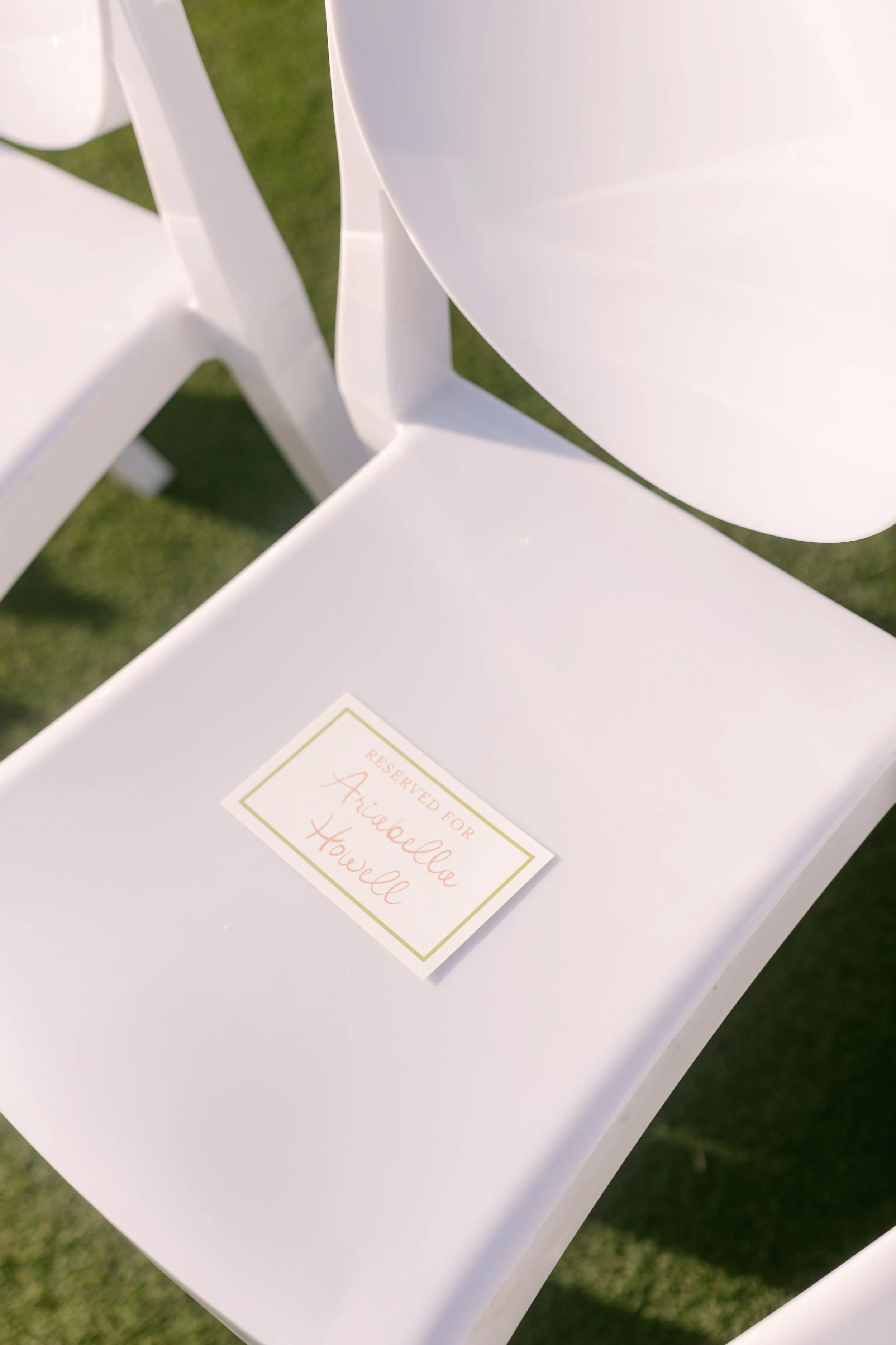 White chair with a reserved sign for Ariabella Howell placed on the seat, outdoors on grass.