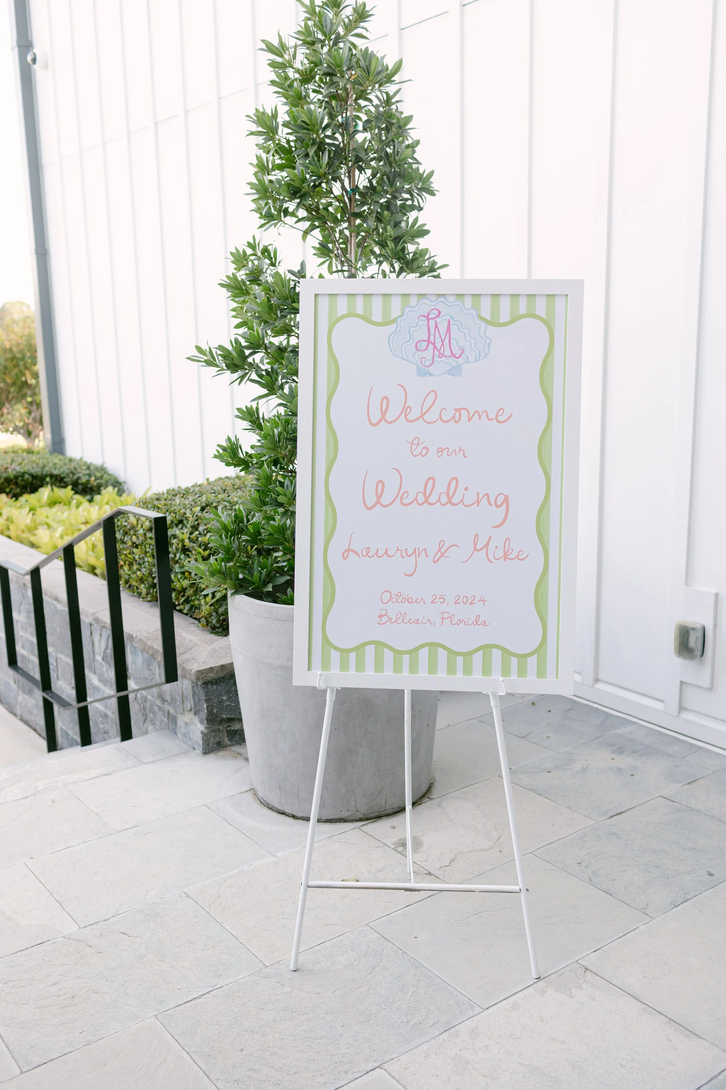 Wedding welcome sign on a stand with green striped border, greeting guests for Lauryn and Mike's wedding on October 25, 2024, in Belleair, Florida, next to a potted plant.