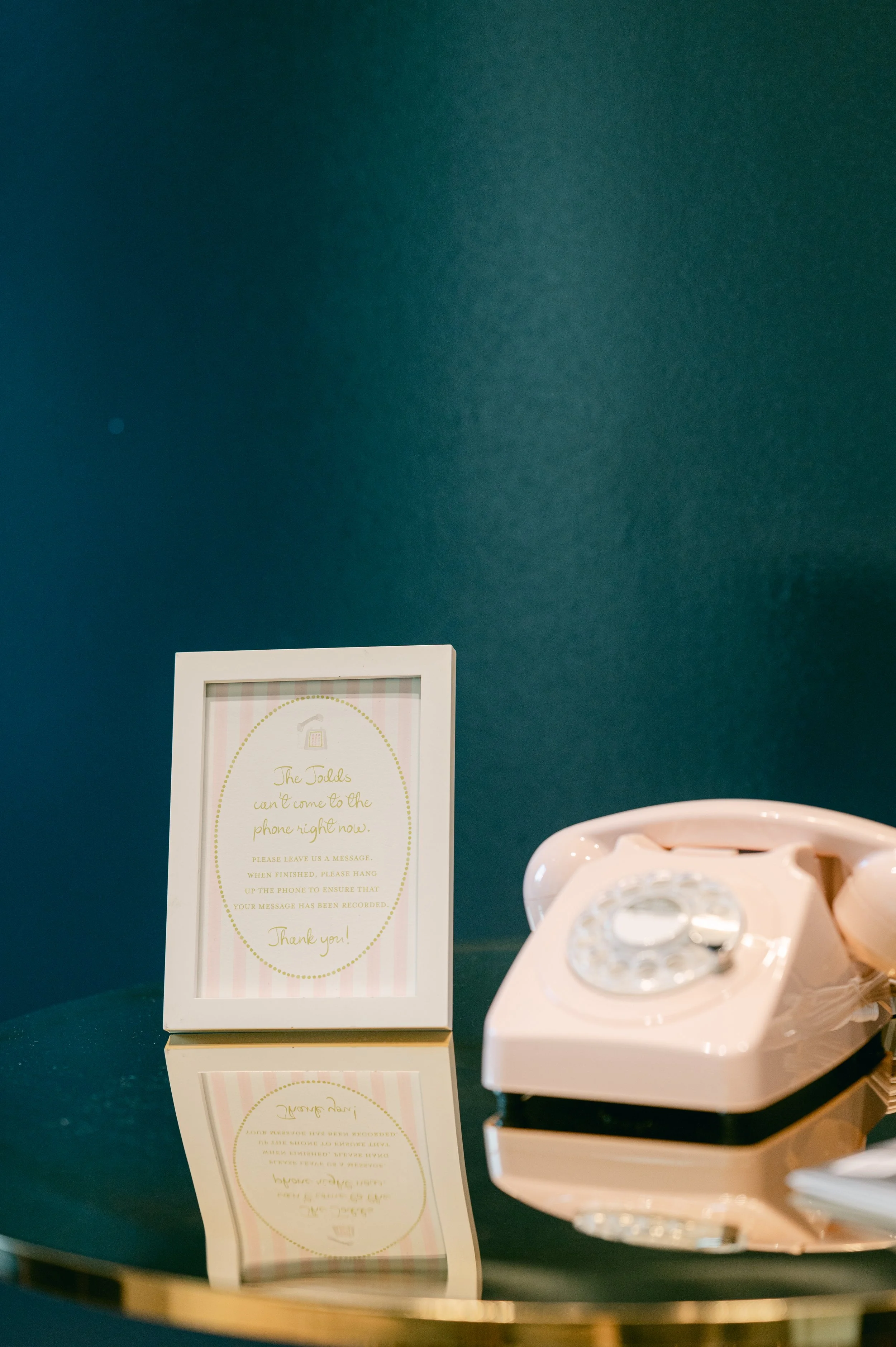 A vintage rotary telephone in cream color and a framed sign with instructions for leaving a message, placed on a reflective surface against a dark teal background.