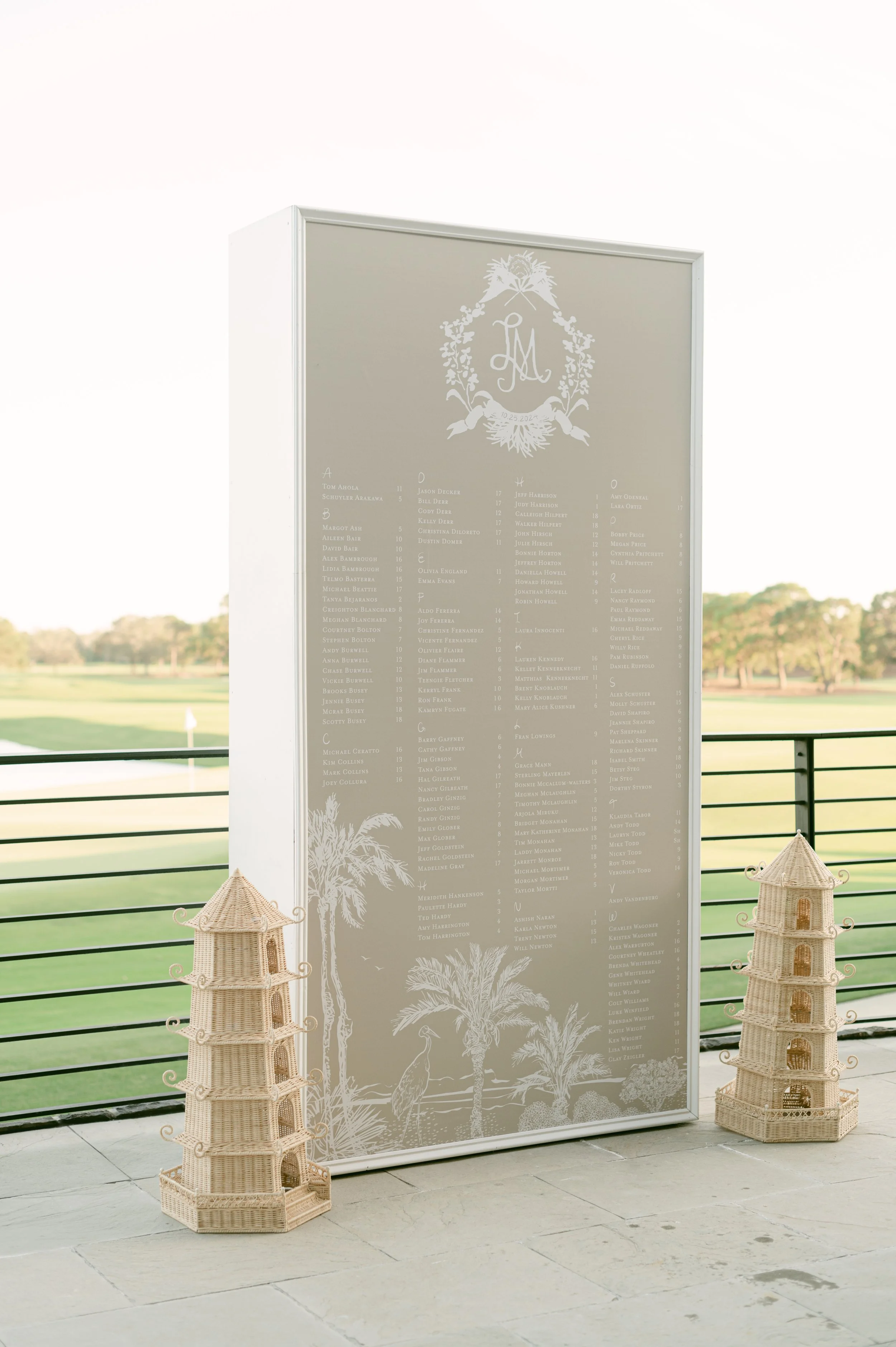 A large wedding seating chart displayed on a stand, with two wicker pagoda-shaped decorations on either side, set against an outdoor view of a golf course with trees and a flag.