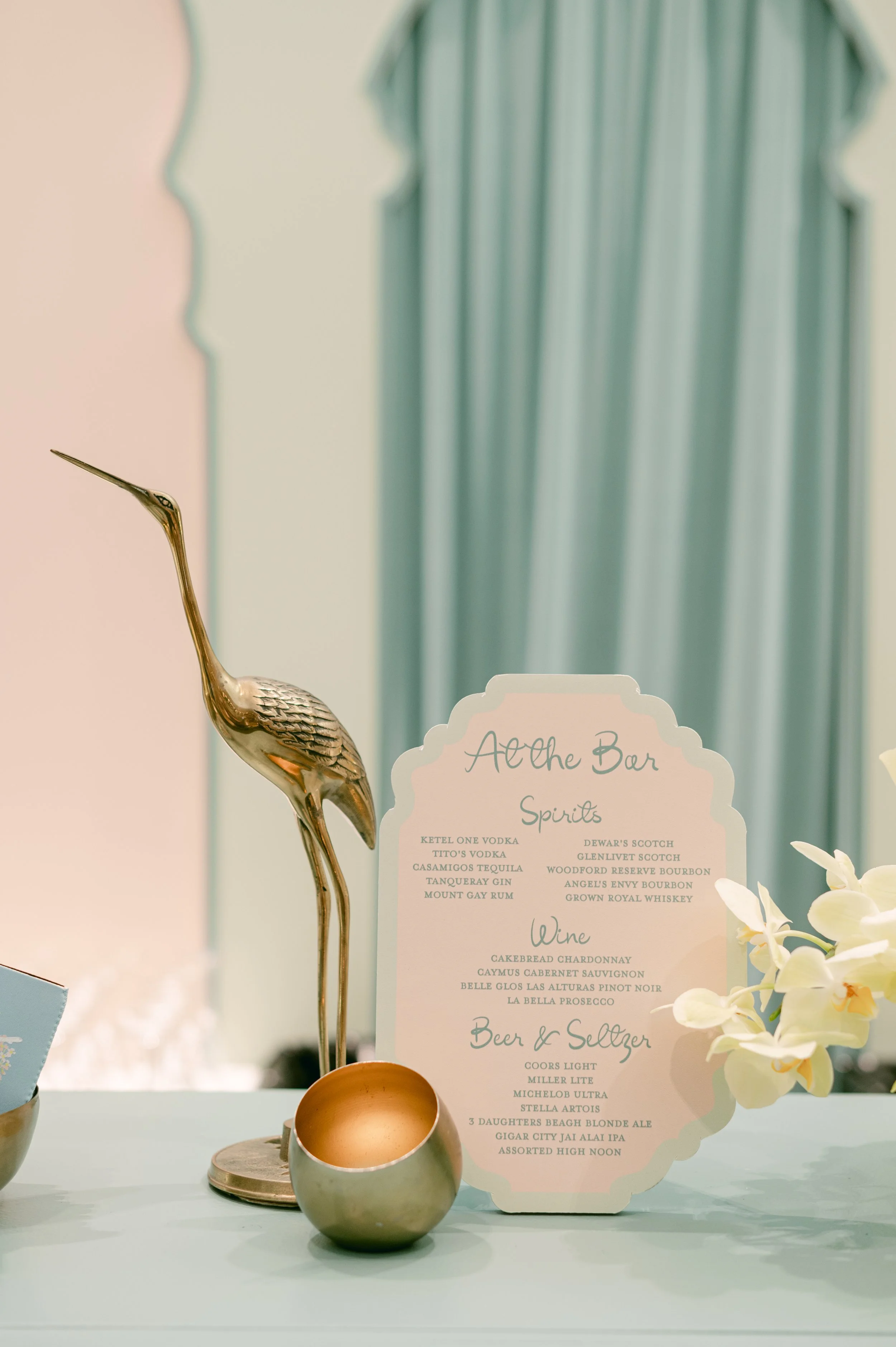 A beverage menu on a table with a bronze-colored bird figurine, white orchids, and a candle, with a pastel-colored background and teal curtains.